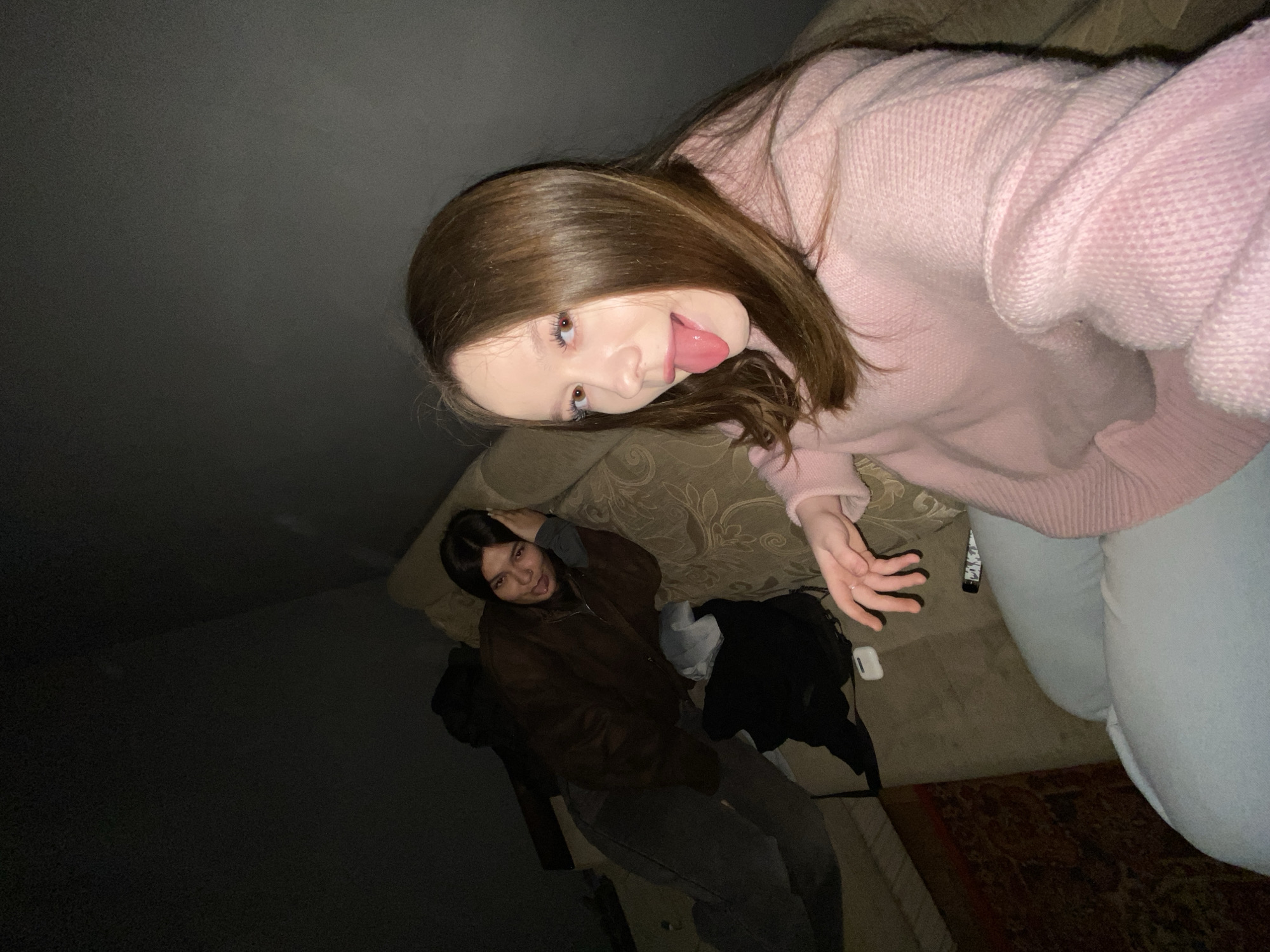 Two young women are situated in an indoor, dimly lit location. The woman in the foreground wears a pink sweater and light-colored jeans, and she is sticking out her tongue playfully. She is positioned in a way that suggests she might be on a couch. Behind her, another woman is lying on the couch, wearing a brown jacket and grey pants, with her hand on her head. A black backpack and possibly a white case are near the woman on the couch. Part of an area rug is visible in the lower right. The image is angled, and the environment is dark, especially toward the left side of the frame. There is no definitive information in the image to confirm the location as Chaikovsky, Russia.