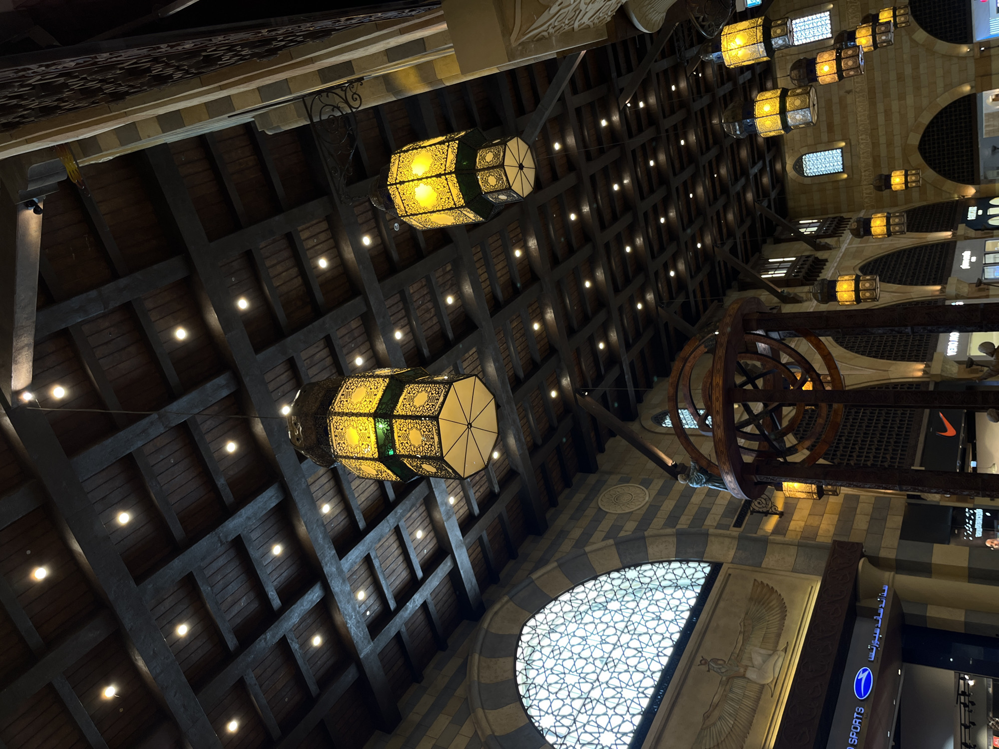 The image captures a view looking upwards at the interior architecture of the Souk Madinat Jumeirah in Dubai, United Arab Emirates. The scene is dominated by a richly detailed wooden coffered ceiling with numerous recessed lights. Suspended from the ceiling are several ornate, multi-tiered lanterns, primarily in shades of yellow and green, casting a warm glow.
In the foreground, a large, circular, wooden decorative structure, possibly a model of an astrolabe or similar astronomical device, is partially visible. To the left, a large arched window with intricate geometric patterned glass is prominent. Below this window, there are shopfronts with illuminated signs, one clearly displaying "SPORTS" and another with Arabic text, indicating retail activity. An emblem featuring a winged figure, reminiscent of ancient Egyptian art, is depicted on the wall above the shopfronts.
Further into the background, more architectural elements are visible, including arched doorways, windows with traditional latticework, and stone construction. The overall impression is one of a vibrant marketplace with a blend of traditional Arabic and contemporary design elements, illuminated by decorative lighting. No specific individuals are clearly identifiable or engaged in any notable actions. The environment suggests an indoor public space, likely a shopping complex or a cultural hub designed to evoke a historical souk atmosphere.