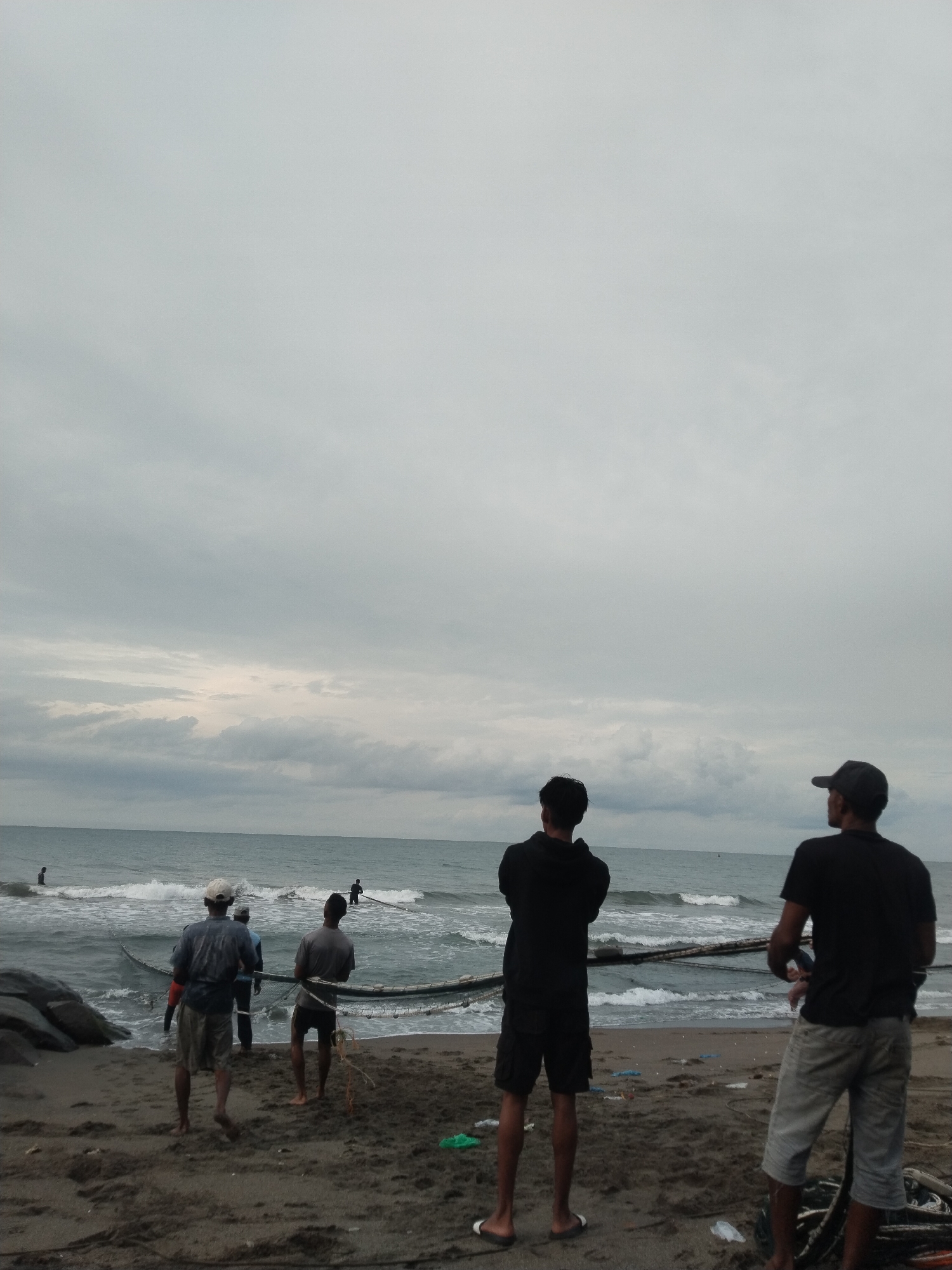 A group of men are engaged in or observing traditional net fishing on a beach in Lhokseumawe, Indonesia, under an overcast sky. In the foreground, two men stand with their backs to the viewer, observing the scene. The man on the left wears a black hoodie and shorts, while the man on the right wears a dark t-shirt, light shorts, and a baseball cap.
Closer to the water's edge, three other men are actively pulling a large fishing net from the ocean onto the sandy shore. The net forms a wide arc in the water, characteristic of seine fishing. Additional figures are visible further out in the water, suggesting more individuals involved in deploying or maneuvering the net.
The scene depicts a sandy beach with dark rocks on the left side. The ocean has moderate waves breaking towards the shore. The sky is heavily overcast with no direct sunlight visible, indicating a cloudy day, possibly during early morning or late afternoon with diffused light. There is some scattered debris visible on the sand. No text is visible in the image.