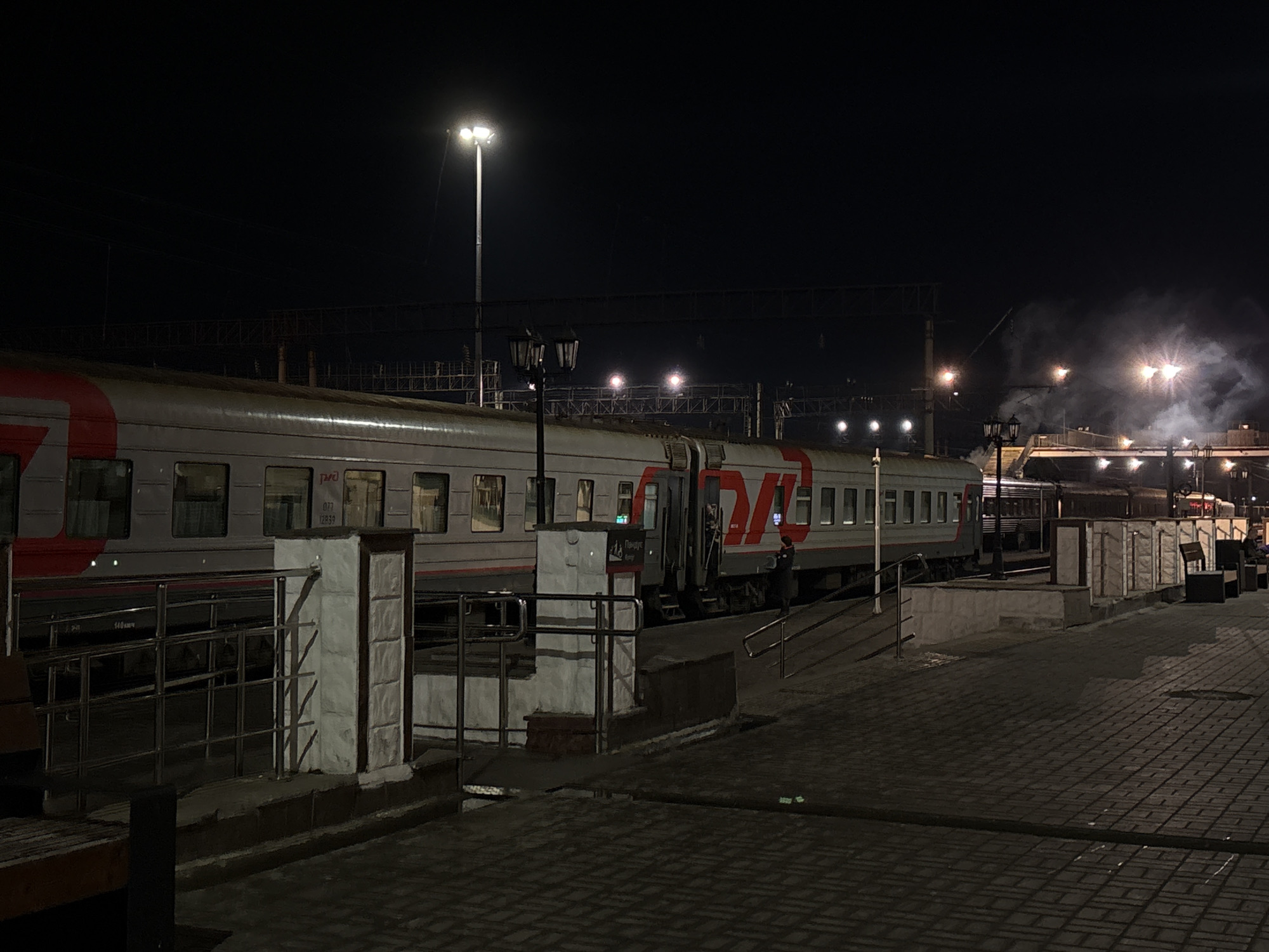 The image depicts a train station at night, likely in Izhevsk, Russia, characterized by the presence of RZD (Russian Railways) passenger train carriages. A train is positioned along a platform, illuminated by overhead lights and street lamps. One person is visible near the train cars. The platform is paved with bricks. Above the station, smoke billows in the distance.