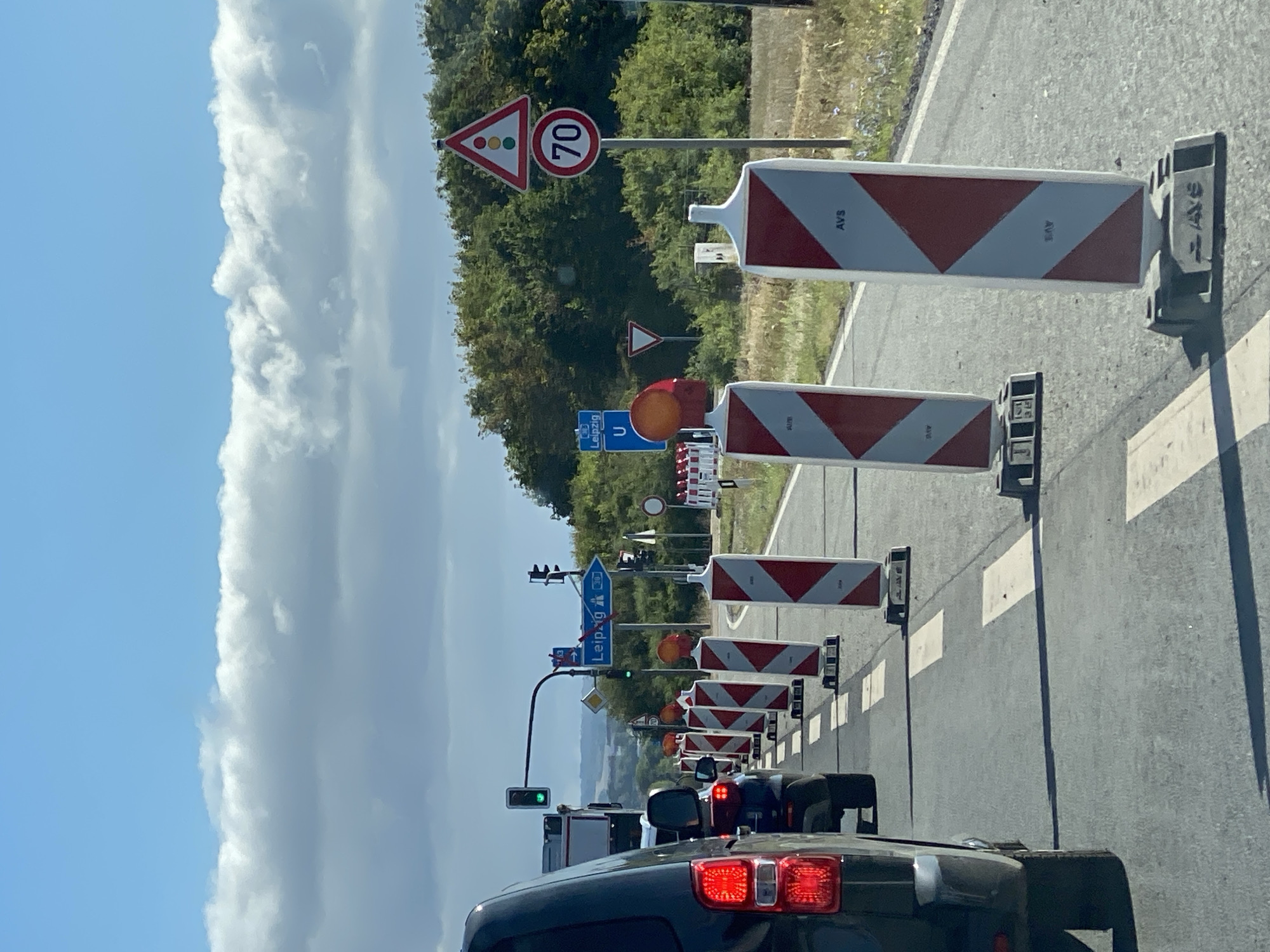 The image depicts a road in Werther, Germany, with traffic cones and signage indicating road construction or a detour. The scene is captured from the perspective of a vehicle, showing the back of another vehicle in the foreground. Several traffic lights are visible, with one showing a green light. A blue road sign points towards "Leipzig". On the right side of the road, a speed limit sign indicates "70", and a triangular warning sign with traffic light symbols is also present. The environment is outdoors, with trees and vegetation on the sides of the road under a partly cloudy sky.