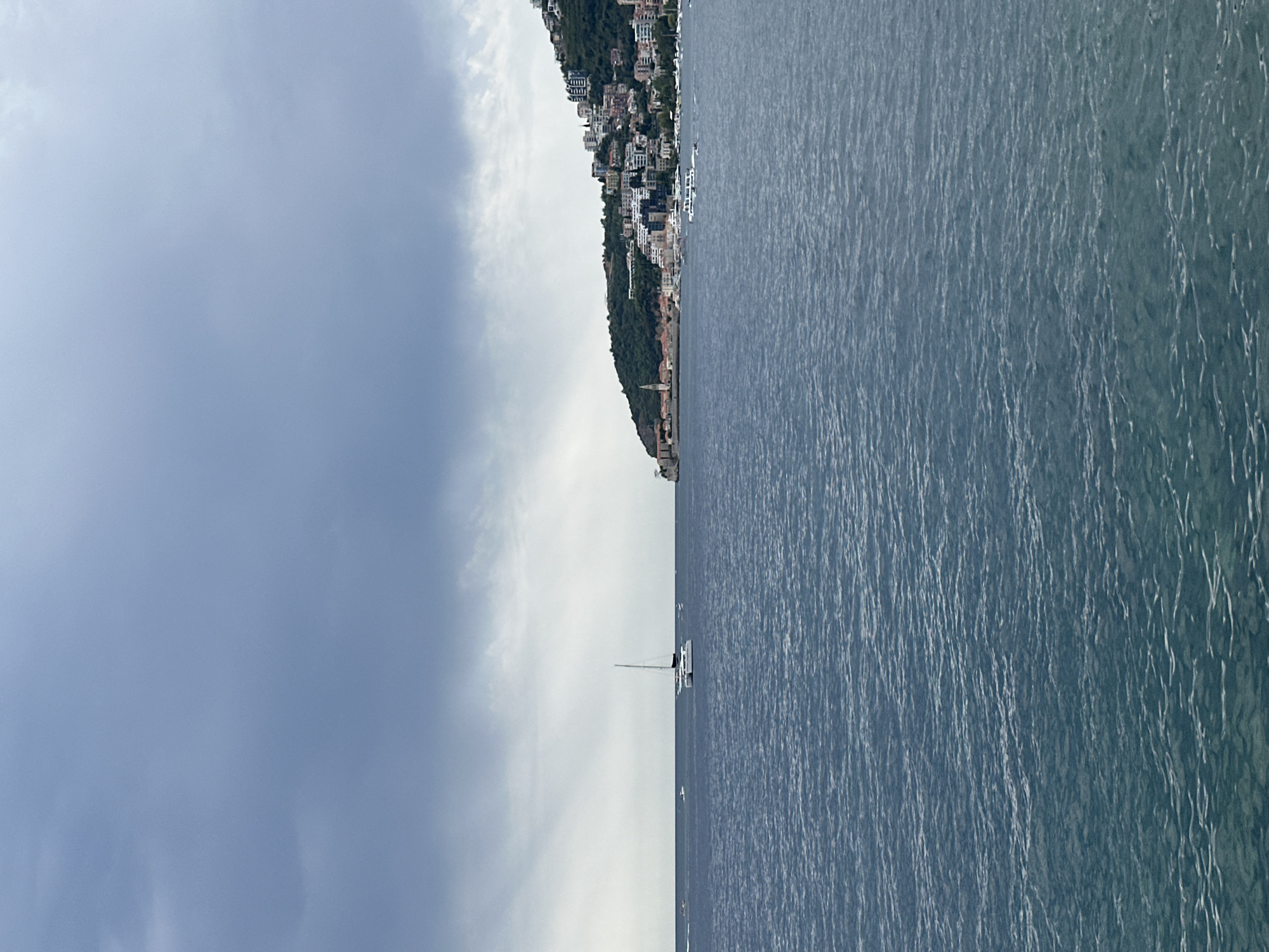 The image shows a wide view of the Adriatic Sea in front of Budva, Montenegro. The water is a deep blue with small ripples. A catamaran is sailing in the middle of the sea, and several other smaller boats are visible in the distance. The coastline of Budva is visible on the right side of the image, with buildings of various sizes and colors climbing up the green hillside. The sky is overcast with dark clouds, suggesting a stormy or cloudy day. The overall impression is one of a coastal city with a busy harbor and a backdrop of natural beauty, under a dramatic sky.