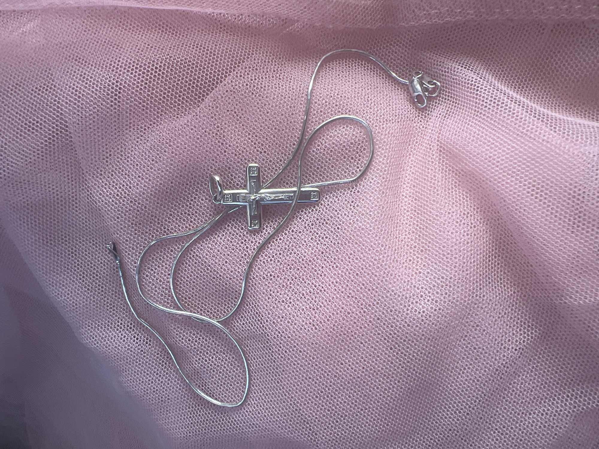 A silver cross pendant necklace is lying on a pink, sheer fabric. The necklace is a thin snake chain with a lobster clasp. The cross pendant is detailed, featuring a depiction of Jesus Christ on it. The fabric has a netted texture and is draped, creating folds and shadows. The scene is lit from above and to the left, casting subtle shadows on the fabric and necklace. There are no people or animals present. The background is a soft focus of the pink fabric. There is no discernible connection to the city of Magnitogorsk, Russia.