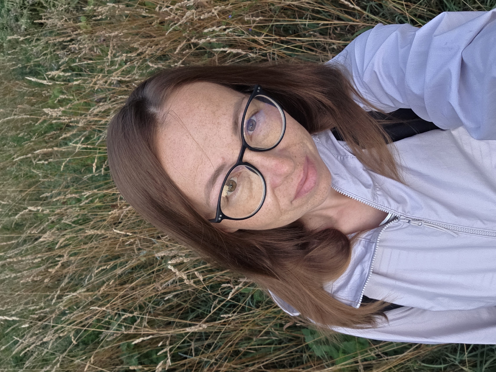 A woman with brown hair and glasses is taking a selfie outdoors. She is wearing a light gray jacket. The background is filled with tall, dry grass, suggesting an autumnal setting. The location is the city of Chaikovsky, Russia.