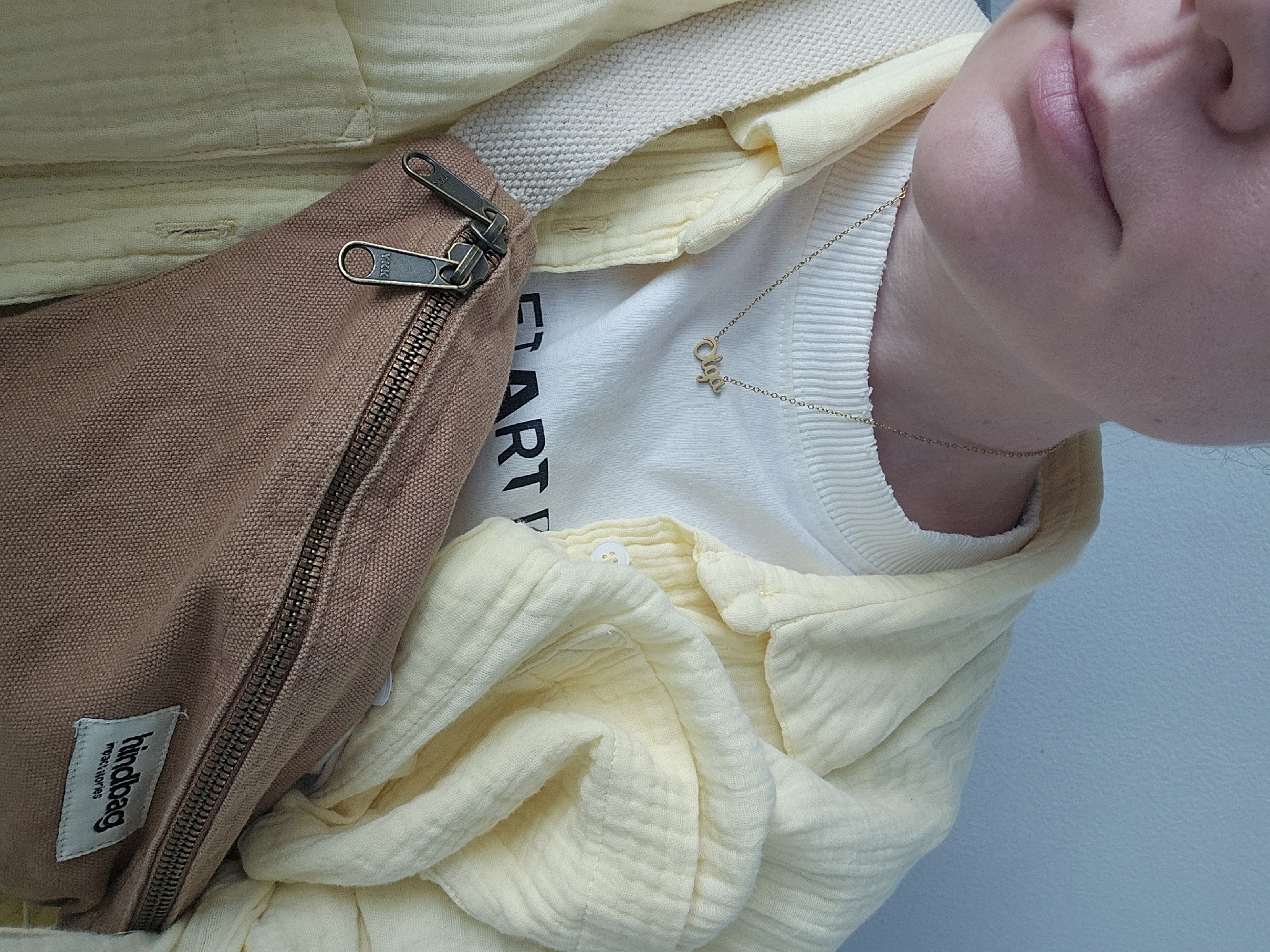 The image shows a close-up view of a person wearing a light yellow, button-up shirt over a white ribbed sweater. A gold necklace with a script initial hangs around their neck. A tan fanny pack with prominent zippers is worn across the chest, with a "hindbag impactstories" tag visible. The background is a plain, light blue wall. There is no indication of the city of Ob, Russia.