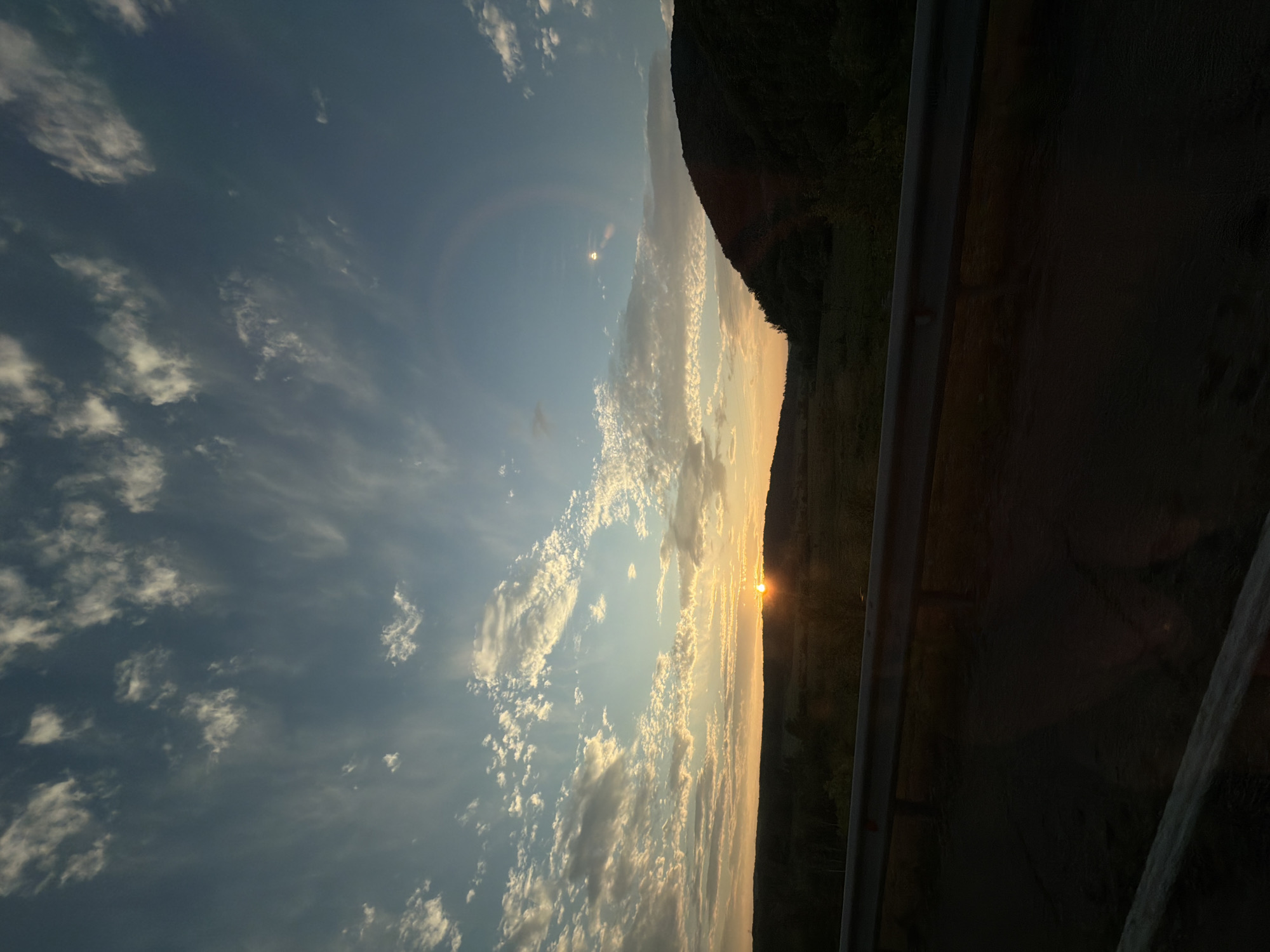 The image captures a sunset over a rural landscape near Birakan, Russia. The sun is low on the horizon, casting a warm orange glow across the sky and illuminating scattered clouds. A dark, tree-covered hill dominates the right side of the frame, while a lower, rolling landscape stretches across the left. In the foreground, a metal guardrail runs along the edge of what appears to be a road, with a dark, asphalt surface visible beneath it. The overall scene is serene and natural, with no people or specific human activity depicted.