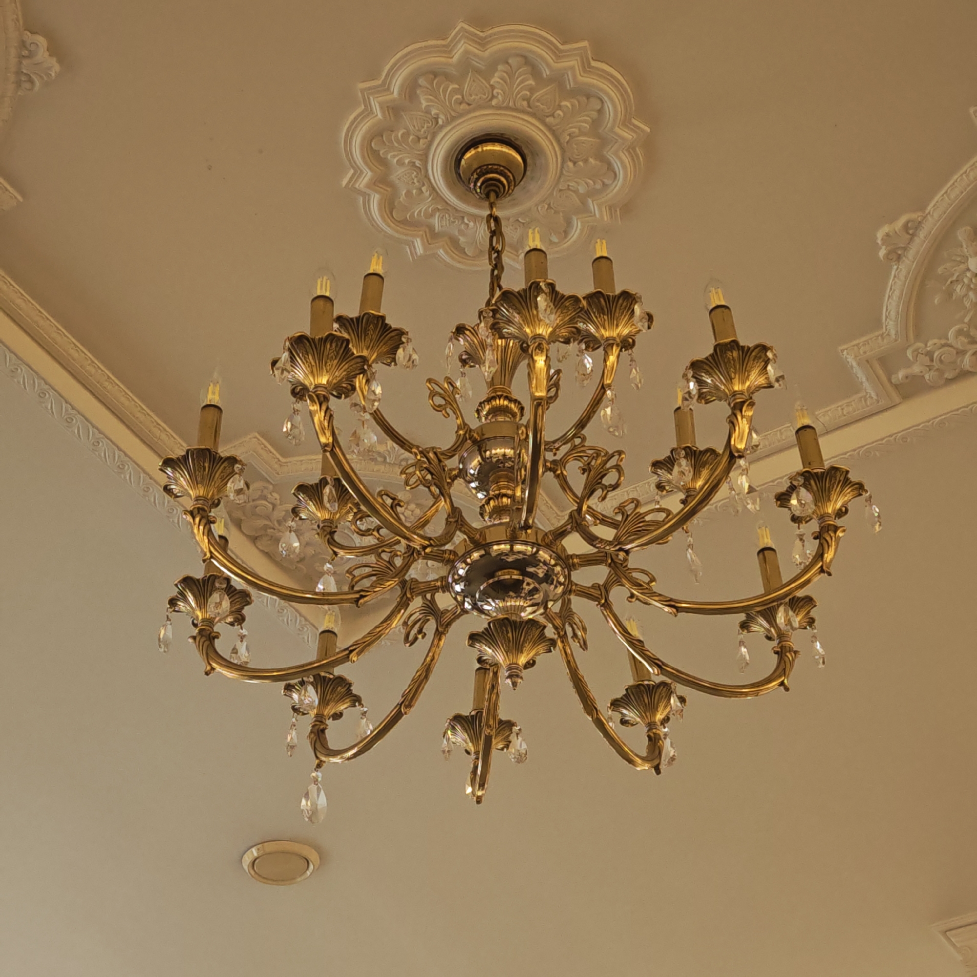 An ornate, multi-armed chandelier dominates the center of the image, viewed from below against a light-colored ceiling. The chandelier is crafted from polished brass or a similar gold-toned metal, featuring numerous gracefully curved arms, each supporting a faux candle-style light bulb and adorned with intricate, leaf-like metallic details. Clear, faceted crystal droplets hang from many points, adding sparkle and reflection.
The chandelier is suspended from an elaborate white plaster ceiling medallion, which features a detailed, lobed design with botanical or classical scroll motifs. Surrounding the central medallion, and extending towards the corners of the ceiling, are additional decorative plaster moldings. These moldings exhibit complex patterns, suggesting a classical or baroque architectural style. The ceiling itself is a plain, light, neutral color, providing a subtle backdrop for the elaborate fixtures.
The scene depicts an interior space, likely a formal room, characterized by its traditional and elegant decor. A small, circular, flush-mounted fixture, possibly another light or a ventilation unit, is visible on the ceiling towards the bottom of the frame. There are no people or specific activities taking place, and no visible text. The overall impression is one of refined grandeur, fitting for a historic or well-appointed building in a location like Ново-Никольское, Russia.