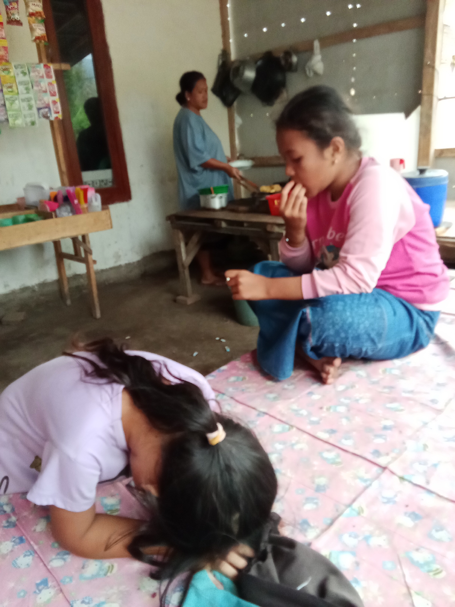 In Trebungan, Indonesia, a cluttered indoor scene depicts domestic activity. Two young females are on the floor, one lying down on a pink mat with a cartoon print, her head resting on her arms, and the other is seated on the mat, eating with her fingers. A woman in a blue shirt stands in the background, appearing to cook or prepare food at a stove. The room has a rustic appearance, with wooden furniture and a simple wall construction. Shelves with various items and what appear to be packets of goods are visible on the left. A large blue container is present on the right. The overall impression is of a humble dwelling where daily life unfolds.