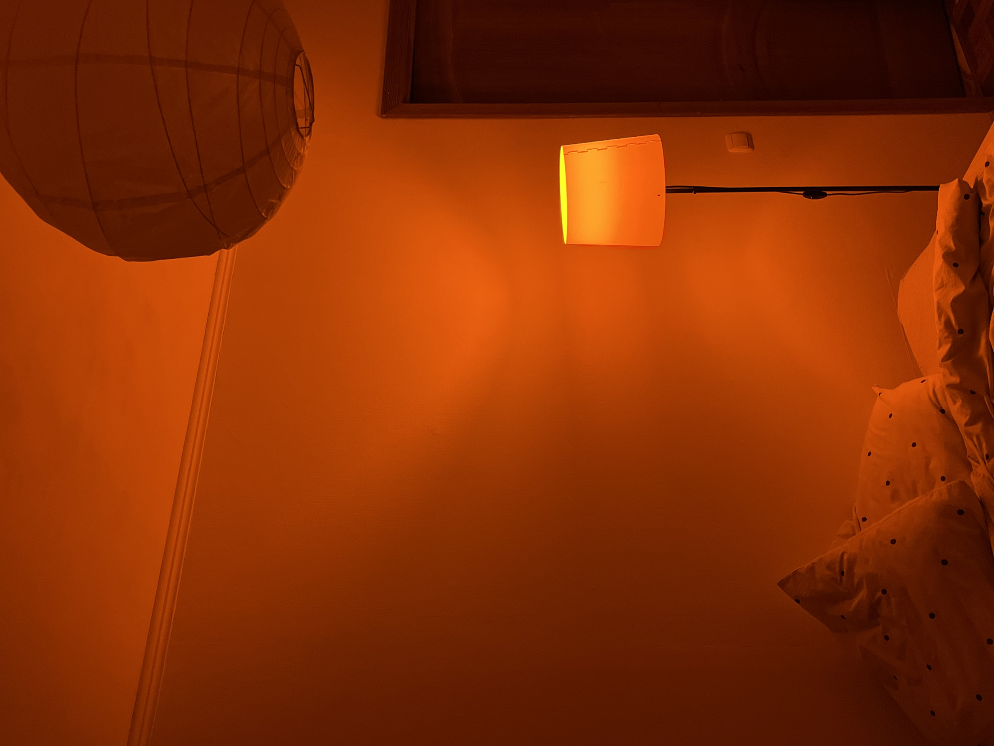 In a room illuminated by an orange glow, a floor lamp stands tall to the right of the frame, its shade casting a warm light on the wall. The lamp has a black metal stand. To the left, a bed is partially visible, adorned with white pillows featuring black polka dots, covered in a duvet. A ceiling light fixture, a paper lantern with a cage-like structure, hangs from the upper right corner of the image. A wooden door stands to the right, slightly ajar. The walls are painted a deep orange, and the ceiling features subtle molding. The floor is not visible. There are no people present.