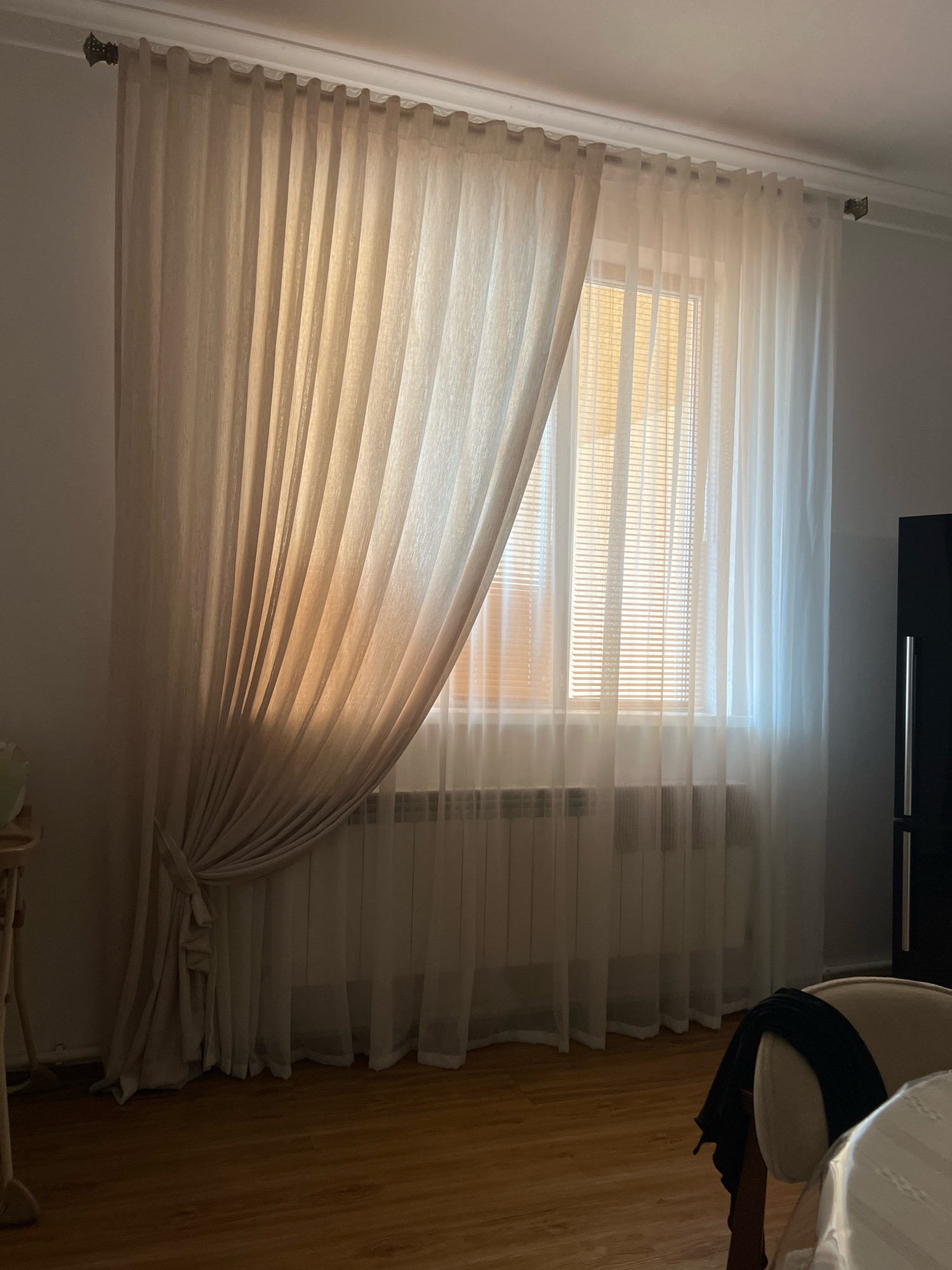 A dimly lit room features a large window covered by sheer beige curtains. The curtains are partially drawn, with the left side draped back and tied with a fabric tie. A set of blinds is visible behind the curtains, with light filtering through. Beneath the window is a white radiator. The floor is wooden and reflects the ambient light. To the right of the window, a dark cabinet or wardrobe is partially visible. In the foreground on the right, a portion of a chair with a dark fabric draped over it is visible. The location is the city of Novogagatli, Russia.