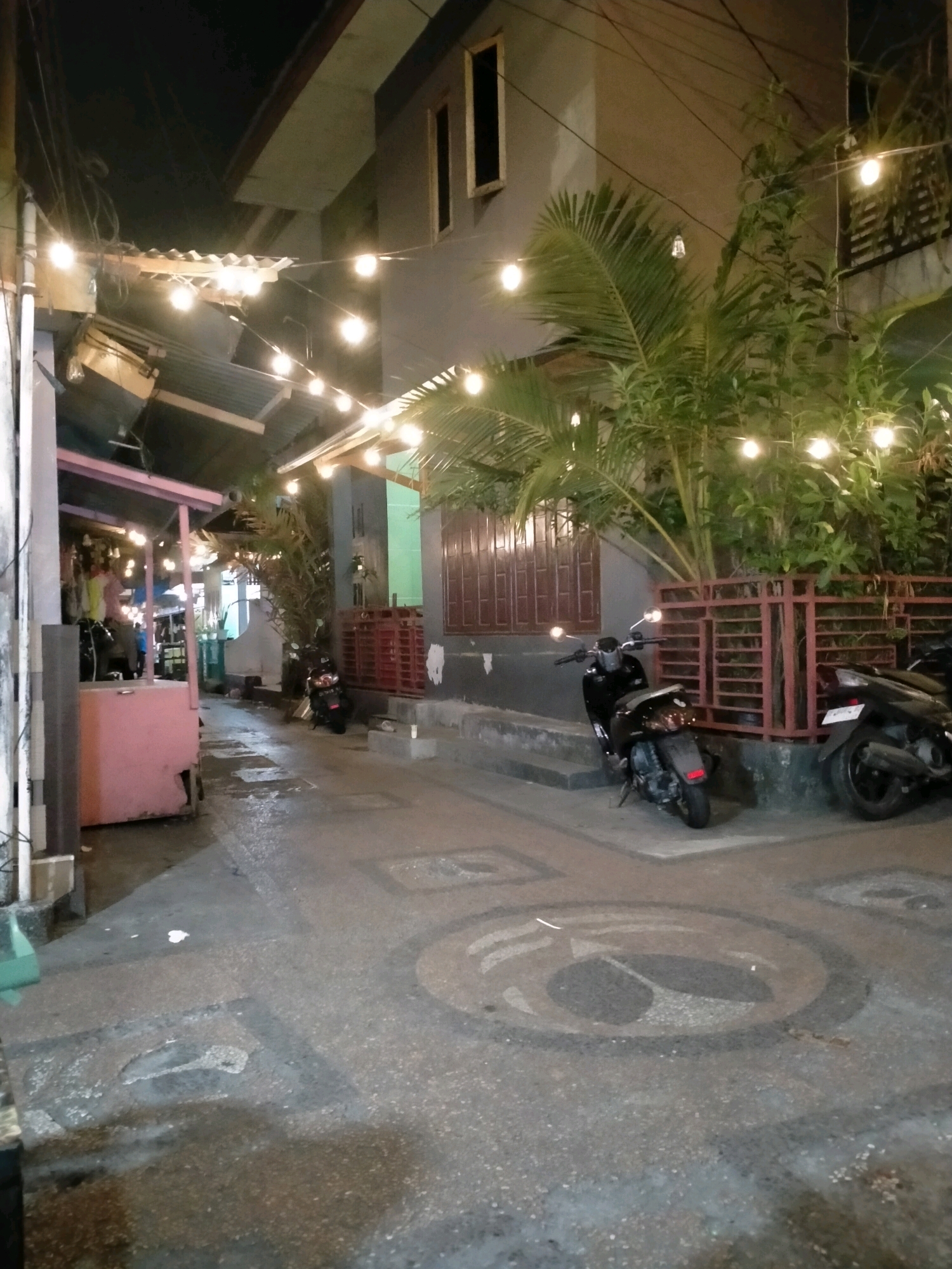 This is a nighttime, eye-level, full shot of a narrow street in Ambon, Indonesia. The street is paved with speckled asphalt featuring circular patterns. To the left, there is a small, pink, open-air food stall with a corrugated metal roof. String lights with exposed bulbs illuminate the scene. In the background, a two-story building with gray walls and dark-paned windows is visible. A red metal fence runs along the front of the building. Two motorcycles are parked on the street: a black one in the foreground and another partially visible to the right. Plants, including palm fronds, grow near the fence. The street appears wet, reflecting the lights. A person is partially visible inside the food stall, and another person is near the stall.