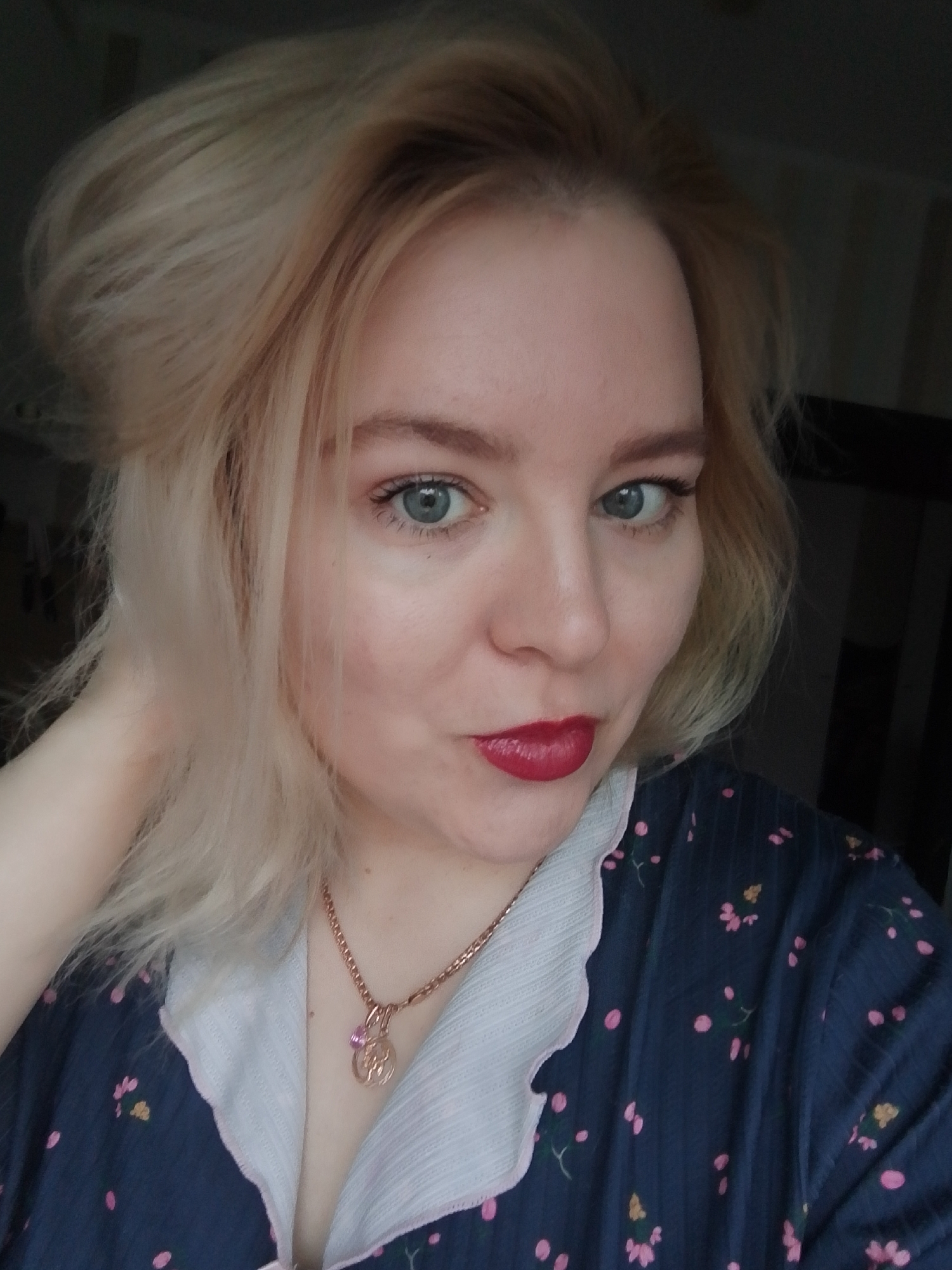 A woman with blonde hair is posing for a selfie. She is wearing a dark blue shirt with pink floral patterns and a white collar. She also has a gold necklace with a pendant. Her lips are painted a glossy red, and her eyes are light blue. The background is blurry, but it appears to be an indoor setting. There is no information to indicate a location in Izhevsk, Russia.