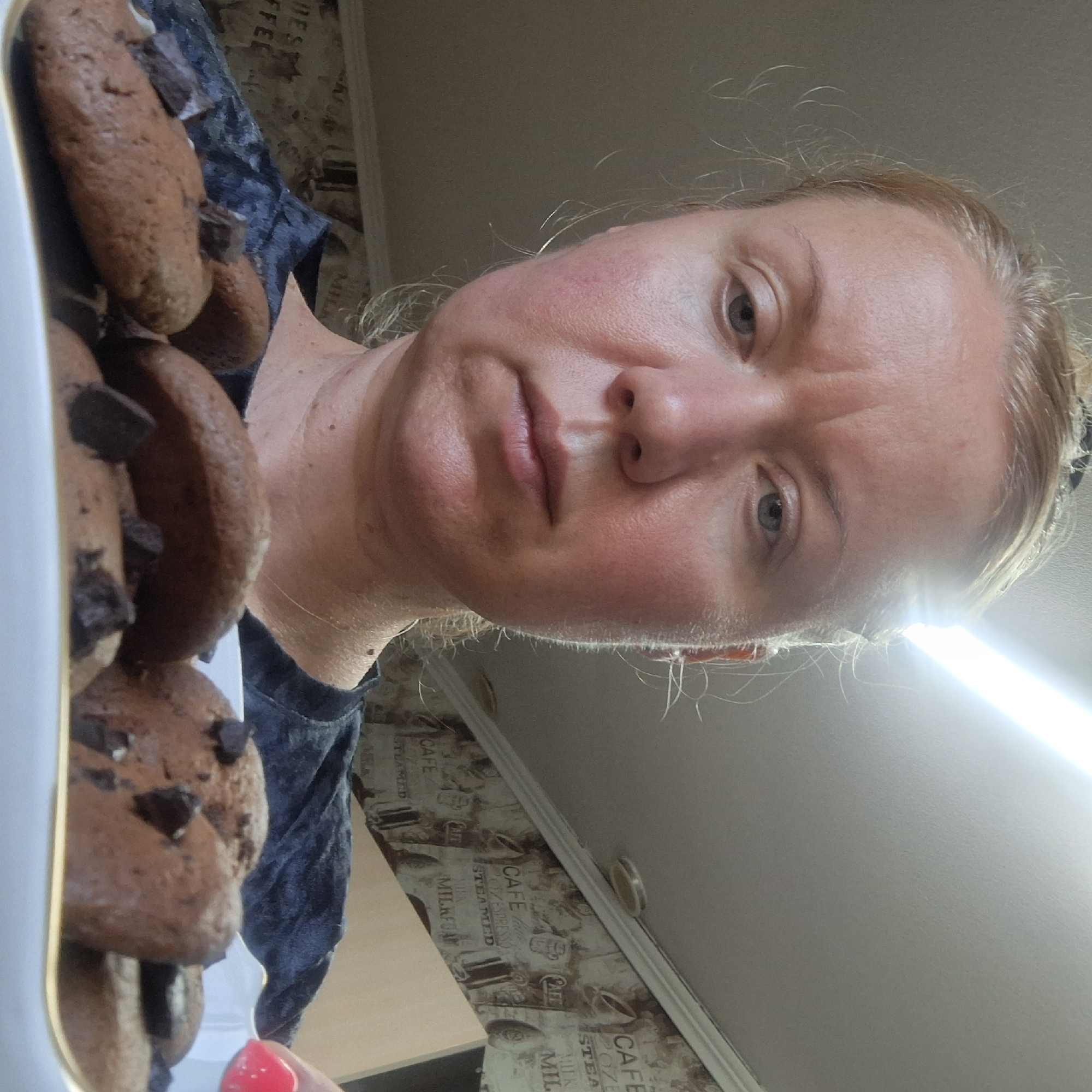 A woman with blonde hair tied in a bun is holding a white plate of chocolate chip cookies. She is wearing a dark blue shirt. The background shows a wall with cafe-themed wallpaper and a bright light fixture on the ceiling. The cookies are brown with dark chocolate chips. The woman's facial expression is neutral.