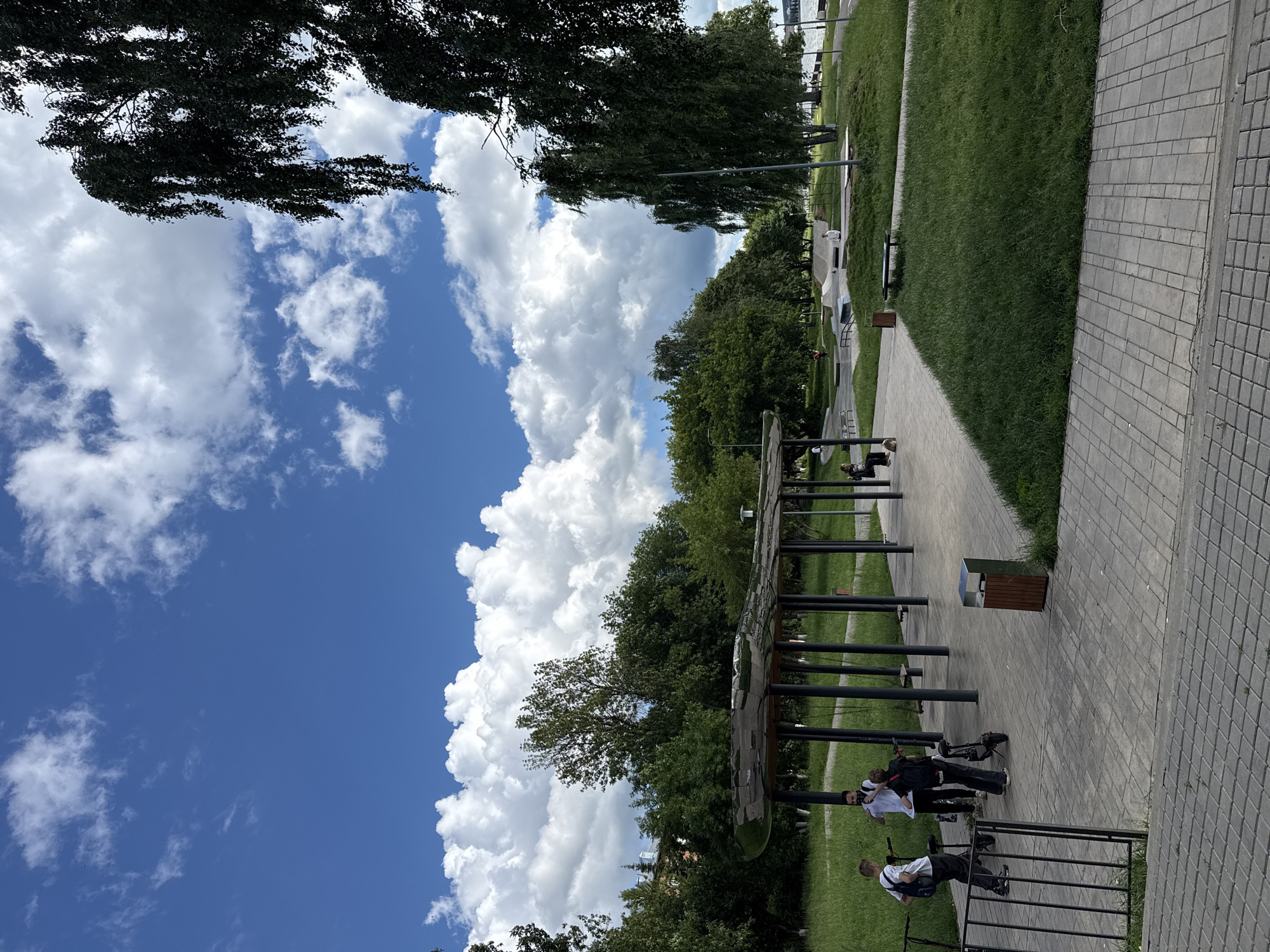 A bright, cloudy day is depicted in this image of a park. The sky is a vibrant blue with large, fluffy white clouds. In the foreground, there is a paved walkway made of grey cobblestones. To the left of the walkway, a black metal fence encloses a grassy area.
Underneath a modern, open-air shelter with a curved, reflective roof and dark poles, several people are visible. Three young males are standing near electric scooters. One is wearing a white t-shirt and dark pants, another a white t-shirt and dark shorts, and the third, a white t-shirt and black pants. A dark-colored trash bin with a silver top is situated near the shelter.
Beyond the shelter, a large expanse of lush green grass stretches out. This area features a skate park with ramps and obstacles. Further in the background, numerous trees with dark green foliage line the park. A body of water, possibly a lake or river, is visible in the distance, with a distinct shoreline. A large tree with drooping branches is prominent in the right side of the frame, its dark leaves contrasting with the bright sky. The overall setting suggests a public recreational space in the city of Votkinsk, Russia, likely a park designed for leisure activities.