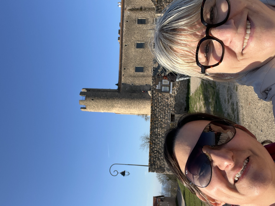 A selfie photograph captures two adult women in the foreground against the backdrop of the Château de Chambles in Chambles, France, under a clear blue sky.
The woman on the left has dark hair, wears dark Ray-Ban sunglasses, and is smiling towards the camera. She is visible from her forehead to her upper chest. The woman on the right has blonde hair, wears tortoiseshell-framed eyeglasses, and is also smiling. She is visible from her forehead to her upper chest.
In the mid-ground, a dirt path leads towards a stone wall. A black decorative street lamp with a scrolled arm and lantern is positioned to the left. A small, light-colored building with a dark roof is partially visible further left. On the stone wall, a sign with dark text on a light background reads "Le Essario." A third individual in dark clothing is partially visible standing near the base of the castle wall to the right of the tower.
In the background, the Château de Chambles stands prominently. It features a tall, round stone tower with crenellated battlements on the left. Adjacent to the tower is a longer, lower stone building with several rectangular windows. The structures are built from light-colored stone. Sparse, bare trees are visible behind the wall and to the left of the castle. A distant landscape is visible on the far left horizon. The sky is bright blue with a faint white contrail visible.