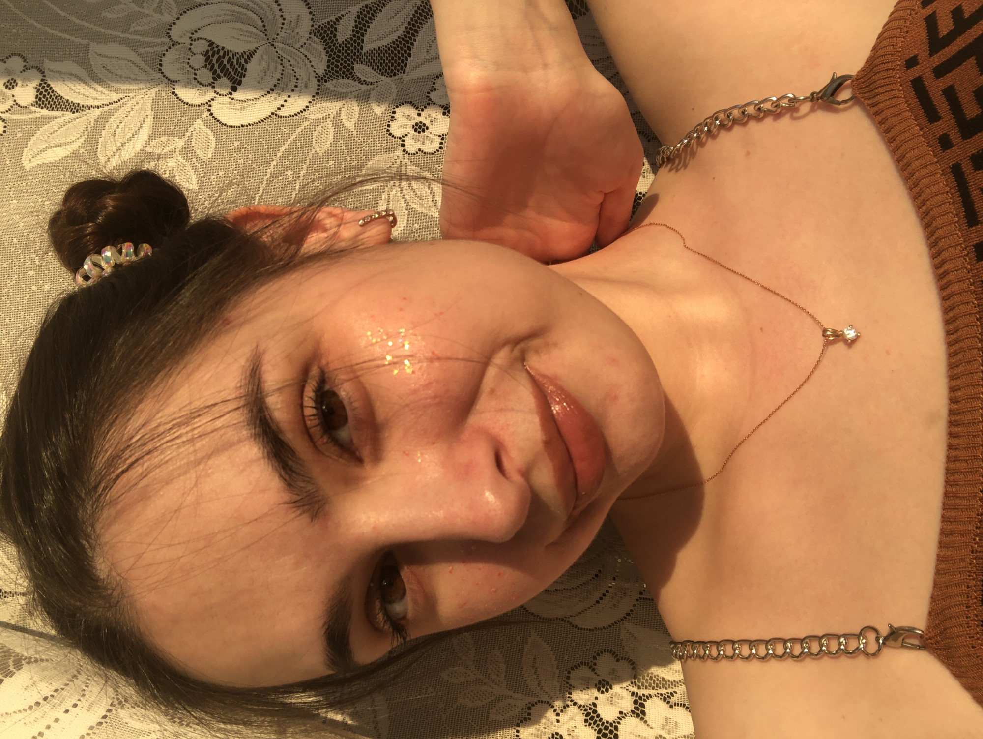 A close-up photograph shows a young woman lying on a floral-patterned surface, possibly a bed or tablecloth. Her head is tilted, and she looks directly at the camera with a slight smile. Her brown hair is styled in a bun. She wears a thin gold necklace with a small diamond pendant. A silver chain adorns her upper arm. A hand rests near her neck. The location, city of Noviy, Russia, cannot be verified from the image.