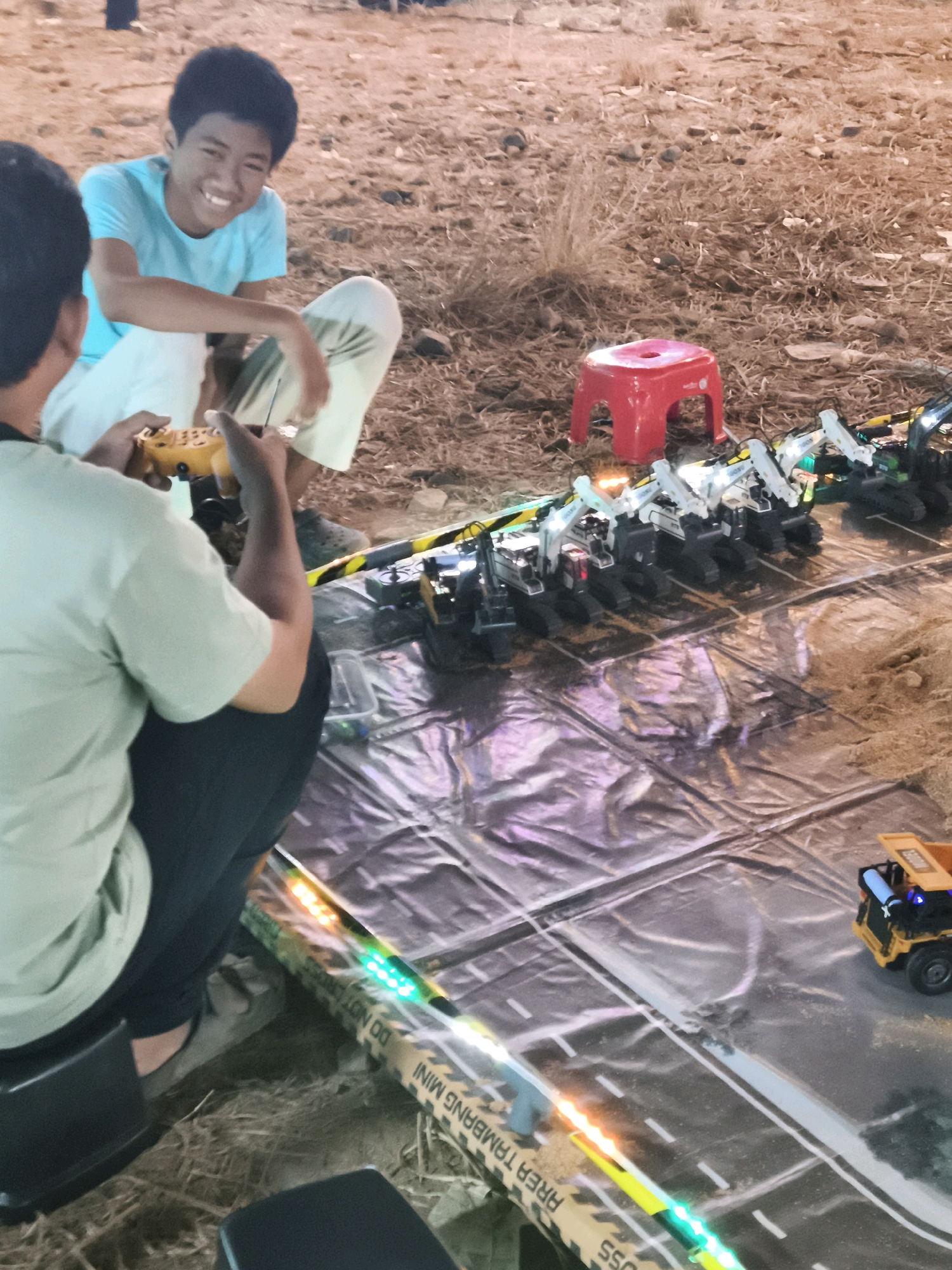 The image depicts a group of young people engaged in an outdoor activity in what appears to be a sandy or dirt area, likely a park or undeveloped space, possibly in the city of Palembang, Indonesia. In the foreground, a person wearing a light green shirt and dark pants is holding a yellow remote control, seemingly operating a toy vehicle. Behind them, another person in a light blue t-shirt and light-colored pants is kneeling and smiling, looking towards the activity.
Spread across a shiny, dark surface, resembling a tarp or a large sheet of plastic, is a miniature road constructed with toy construction vehicles. Several white and black toy excavators are lined up, and a small yellow toy dump truck is visible on the road. The road itself is marked with white lines, simulating traffic lanes. The edge of the play area is bordered by yellow and black caution tape with text that appears to include "AREA TAMBANG MINI" which translates to "MINI MINING AREA" and "HOSS". Colorful LED lights are strung along the edge of the play surface, adding a festive element to the scene.
The background consists of dry, patchy grass, scattered small rocks, and a red stool. The overall impression is of a casual, playful gathering. The lighting suggests it is late afternoon or early evening.