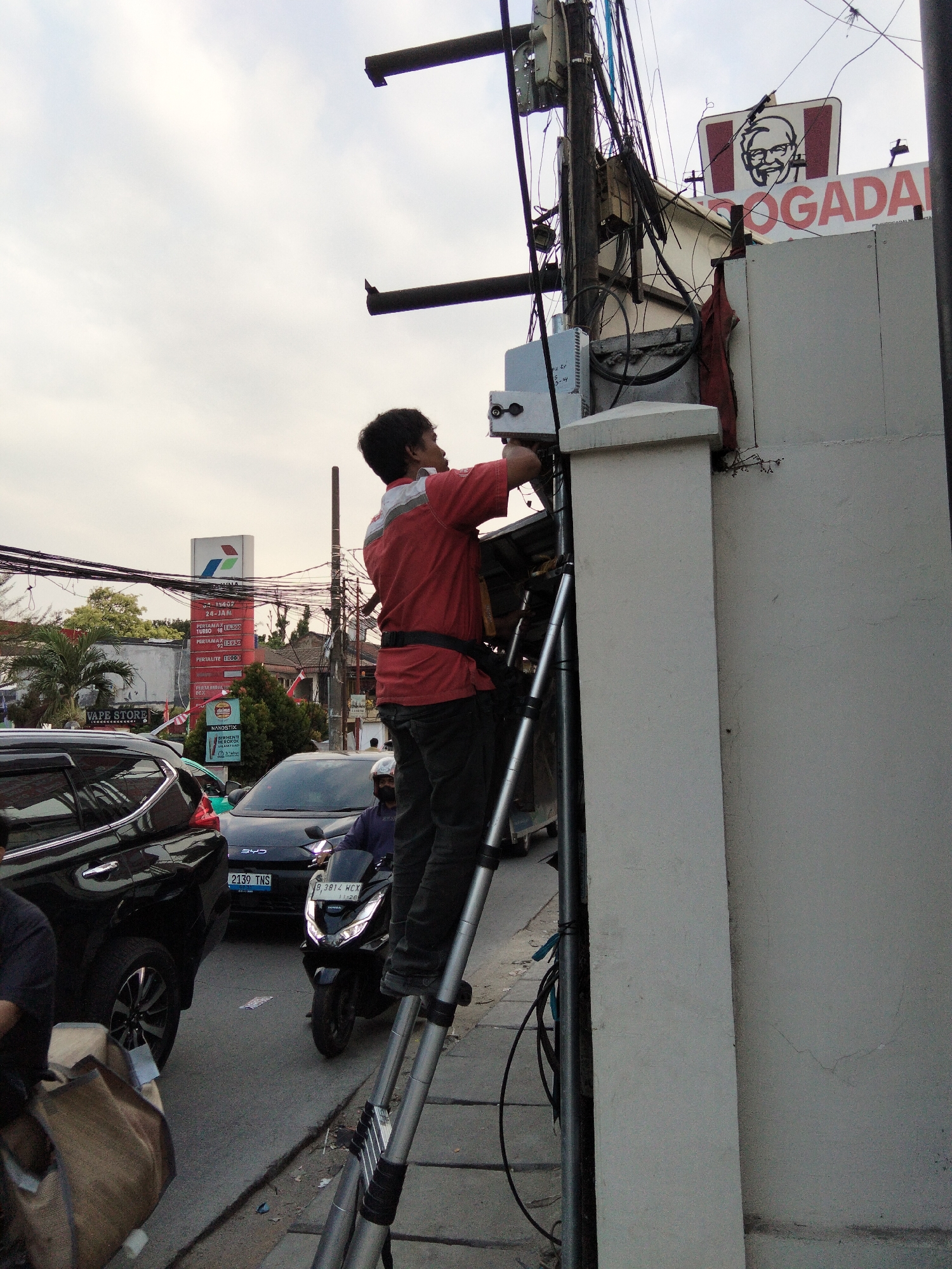 The image depicts a street scene in South Tangerang, Indonesia, likely near a KFC outlet given the visible signage. A man wearing a red shirt and dark pants is on a ladder, working on electrical equipment attached to a utility pole. The pole is laden with numerous wires and cables, indicating a complex network of utilities. In the background, a black SUV and a black scooter are visible on the road, suggesting traffic. A large billboard with the Petroeconomic logo and fuel prices is also present, along with a "Vape Store" sign. The overall environment appears to be an urban street with commercial establishments and a somewhat overcast sky.