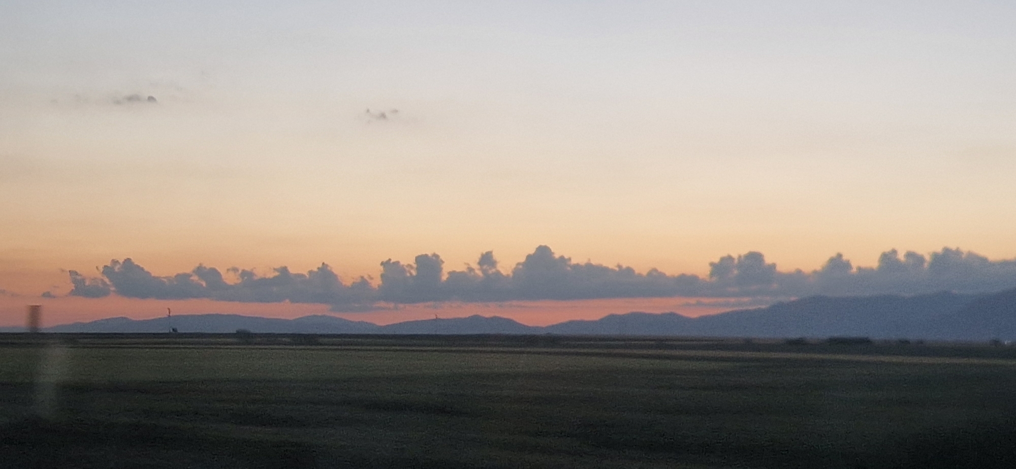 The image shows a landscape at sunset or sunrise. In the foreground is a dark, flat field. Beyond the field, a series of low, rolling hills or mountains are silhouetted against a band of orange and red in the sky. Above the hills, a line of puffy clouds mimics the shape of the mountains below. The sky fades from orange near the horizon to a lighter peach color higher up, with a few sparse clouds visible in the upper portion of the image. The scene is likely rural, and the lighting suggests it's either dawn or dusk. The location and existence of people in the image cannot be verified.