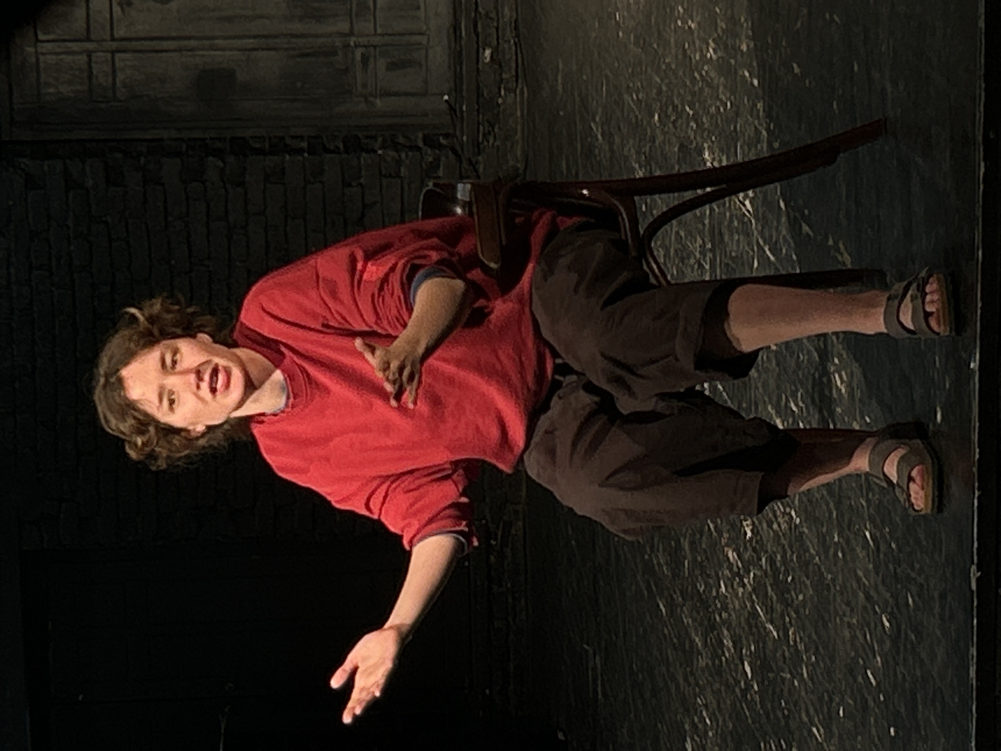 A person is on a stage in what appears to be a play. The stage is dark, with a brick wall visible in the background on one side. The floor is dark with a subtle texture. The person, who has short curly hair, is wearing a red long-sleeved shirt, dark-colored culottes, and sandals. They are positioned to the left of a dark wooden chair and are leaning towards the left with their right arm across their chest and their left arm extended with their hand open. Their face is expressive, with their mouth open as if speaking or acting. The lighting on the stage is focused on the person, leaving the rest of the scene in shadows. The location is Unknown, Unknown.