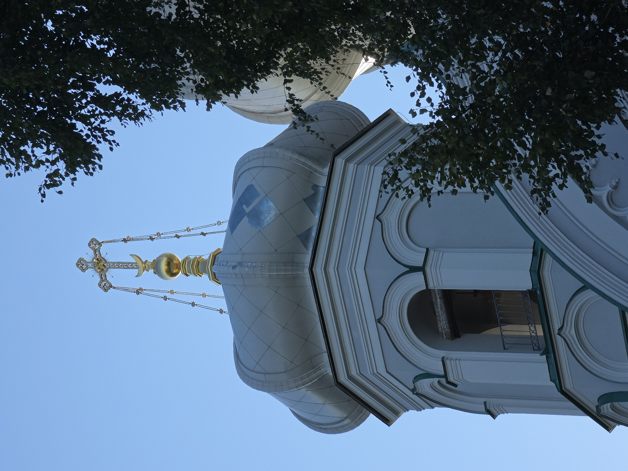 The image shows a close-up of the dome and bell tower of the Pavlovsky Cathedral in Gatchina, Russia. The dome is silver and features a diamond-shaped pattern. Atop the dome is a golden cross and crescent moon, a symbol often found on Orthodox churches. A portion of a leafy green tree is visible on the right side of the image, obscuring part of another dome. The sky is a clear, bright blue. The bell tower has arched openings and decorative trim. No people or animals are visible in the image. The scene is captured from a low angle, looking up at the architectural elements against the sky.