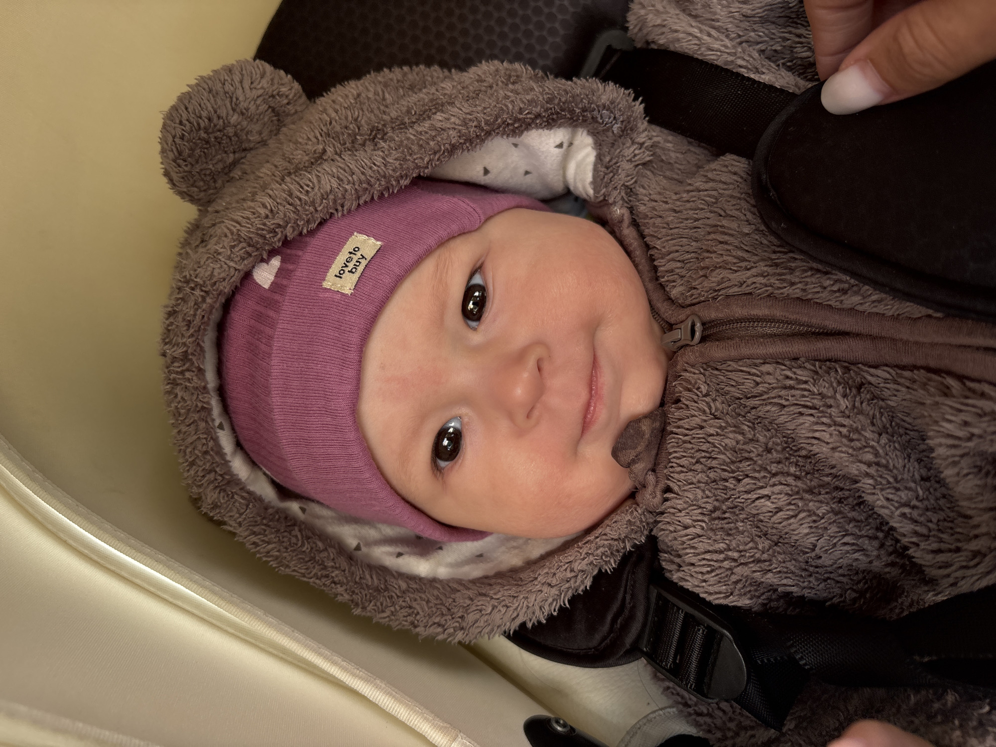 A baby wearing a pink hat and a fuzzy, brown, bear-eared hooded jacket is lying in what appears to be a car seat or stroller. The baby is looking directly at the camera. Part of a hand is visible in the upper right corner of the image. A buckle of a safety harness is seen on the jacket. The location is not identifiable from the image.