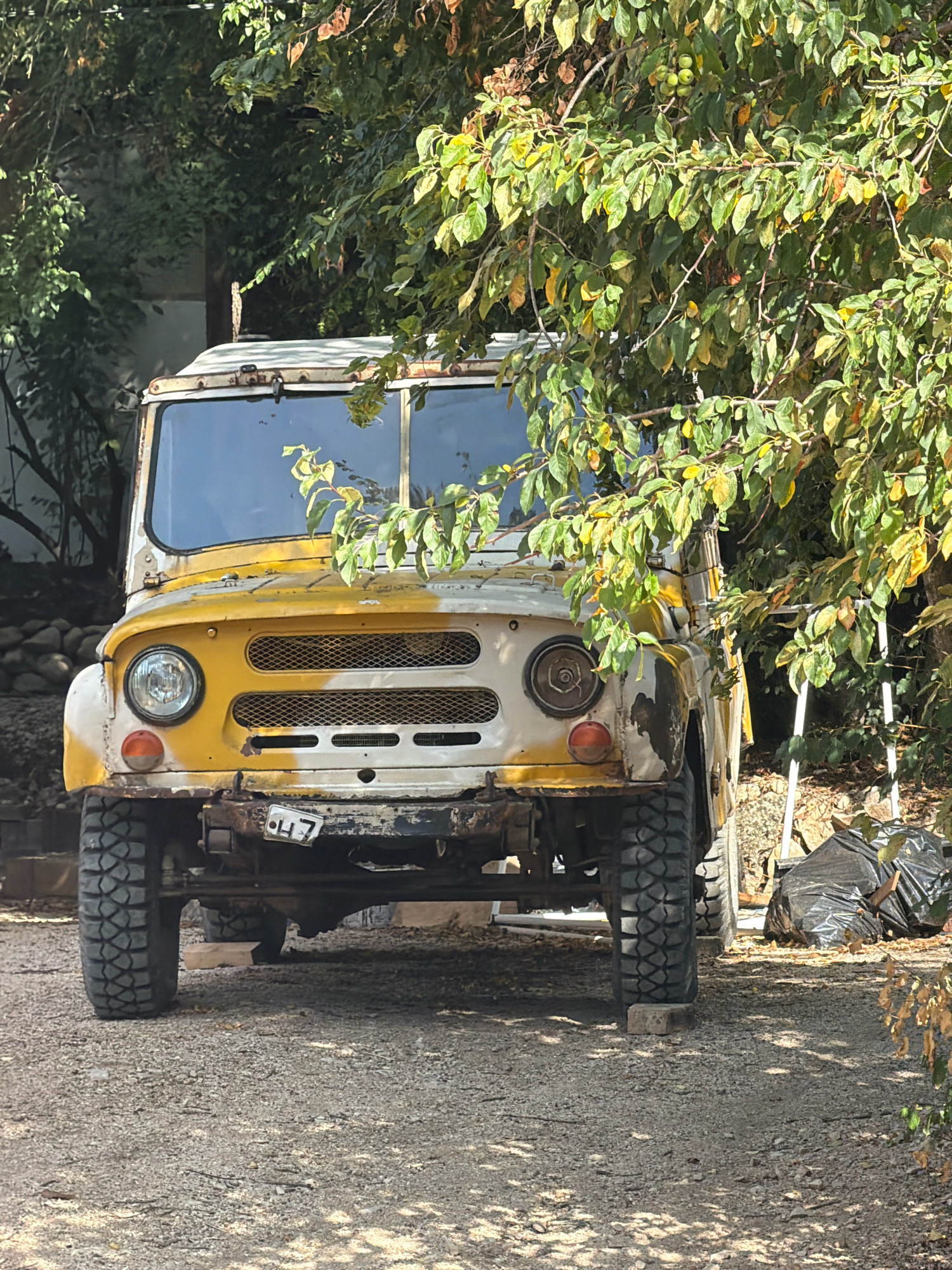 A weathered, yellow and white off-road vehicle, possibly a UAZ-469, is parked on a gravel surface in an outdoor environment. The vehicle shows signs of rust and age, with peeling paint. It is partially obscured by the branches and foliage of a deciduous tree with green and yellowing leaves. The foreground is dominated by the gravel, with some dried leaves scattered around. In the background, a pile of black plastic bags is visible, along with a ladder leaning against what appears to be a structure. The overall scene suggests a rural or semi-abandoned setting, possibly a yard or a field. The lighting indicates it is daytime, with shadows cast by the tree branches on the ground. The location is identified as Balaklavsky District, Ukraine. There are no people visible in the image.
