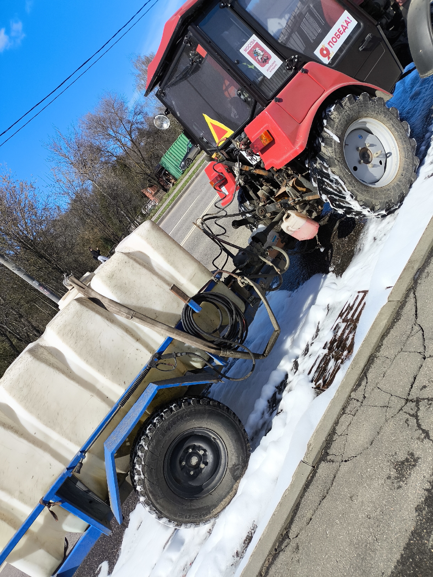 A red tractor pulling a white tank trailer is parked on the side of a road in Moscow, Russia. The tractor has a sticker with the image of St. George slaying the dragon with the word "Moscow" and a sticker of a ribbon with the word "Victory!". The tractor has released a large quantity of white foam, potentially soap, onto the road and over a storm drain. A person is standing on top of the tank trailer. The sky is blue and there are trees without leaves in the background. A green semi-trailer is parked on the roadside across the road from the tractor.
