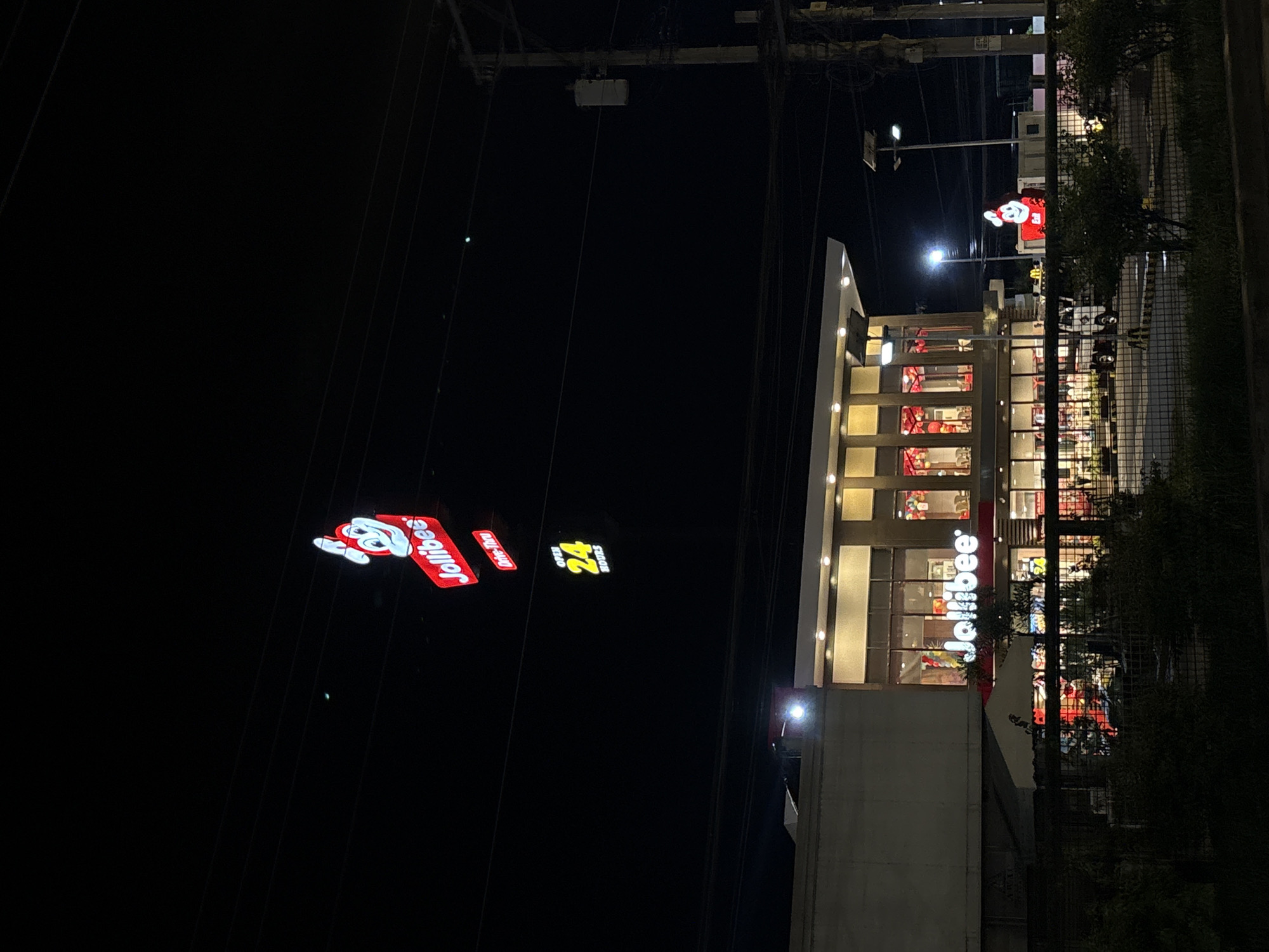 The image shows a Jollibee fast-food restaurant at night in Davao City, Philippines. The restaurant is illuminated by its signage and interior lights. A large red sign with the Jollibee logo and the text "Jollibee" is visible at the top, along with a "Drive-Thru" sign. Below that, a yellow sign indicates "Open 24 Hours". The building has a modern facade with large windows displaying festive decorations, likely balloons. In the foreground, there is a fence and some vegetation. Several vehicles, including a white delivery truck, are parked in the restaurant's lot. The scene is characteristic of a commercial area in Davao City, with utility poles and overhead wires visible in the background, indicating an urban environment.