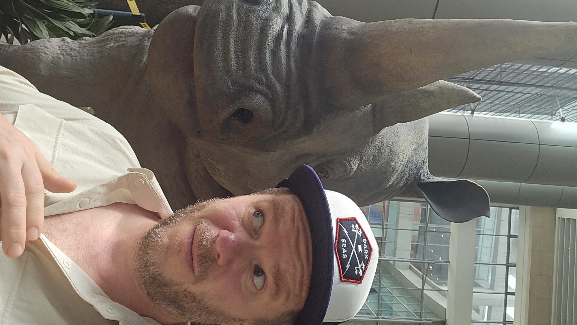 The image shows a man wearing a light-colored button-up shirt and a white and navy baseball cap with a "DARK SEAS" logo. He is standing next to a large rhino statue. The man is looking to his right, with his left hand touching his chest. In the background, there are modern architectural elements, including glass windows and large cylindrical pillars. The setting appears to be an indoor public space, possibly a museum or a shopping mall, given the scale of the statue and the architecture. The overall lighting suggests an interior environment. The image composition is a close-up selfie, with the man's face and the rhino's head prominently featured. The image quality is moderately clear, with some graininess.