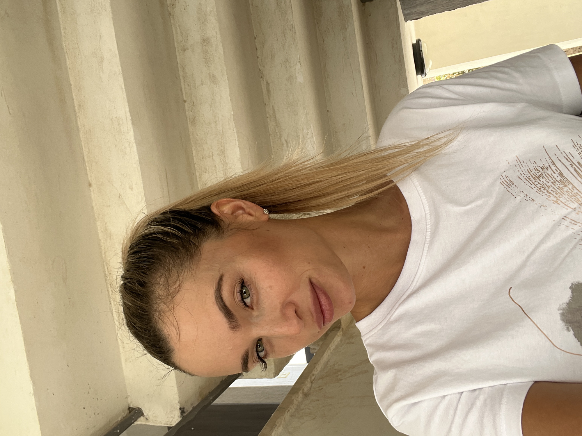 A woman with blonde hair in a ponytail and wearing a white t-shirt stands in front of a concrete staircase. She is looking at the camera with green eyes and a slight smile. She is wearing a silver earring in her left ear. The scene appears to be outdoors, possibly in a courtyard or an urban setting with exposed concrete structures.