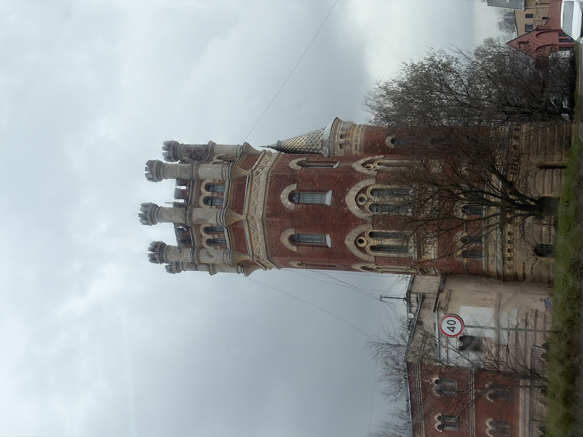 The image shows the Water Tower of the former St. George Community of Sisters of Mercy in Saint Petersburg, Russia, under an overcast sky. The tower, constructed of red brick with light-colored architectural details, features crenellated turrets and arched windows. Adjacent to the tower is a building with a speed limit sign displaying "40" in a red circle. Bare trees are visible around the buildings. The scene is viewed from ground level. The architecture is predominantly Gothic Revival style.