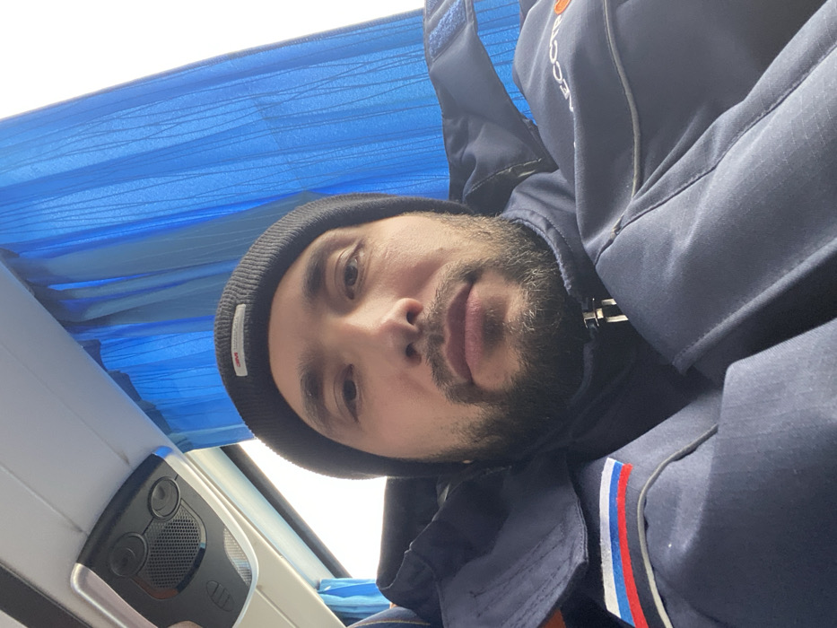 A male individual is depicted in a medium shot, facing directly forward, presumably taking a selfie. He possesses a beard and mustache, dark eyes, and a neutral facial expression. The man wears a black knitted beanie with a white rectangular "3M Thinsulate" label on the front. He is dressed in a dark blue, possibly waterproof or insulated, jacket with a zippered front closure. On the left upper arm or shoulder of the jacket, a patch with horizontal white, blue, and red stripes, resembling the flag of Russia, is visible. A faint, partially obscured logo, appearing orange and beginning with Cyrillic characters like "АЕ ССТР," is on the right chest/shoulder area of his jacket. The environment is the interior of what appears to be a passenger vehicle, likely a bus or coach, characterized by blue pleated curtains covering a window to the right of the subject and a light gray overhead panel with circular vents/lights and a speaker grille positioned above and to the left. The window reveals a bright, overcast sky. The specific location within the city of Luzhitsy, Russia, cannot be determined from visual cues within the image.