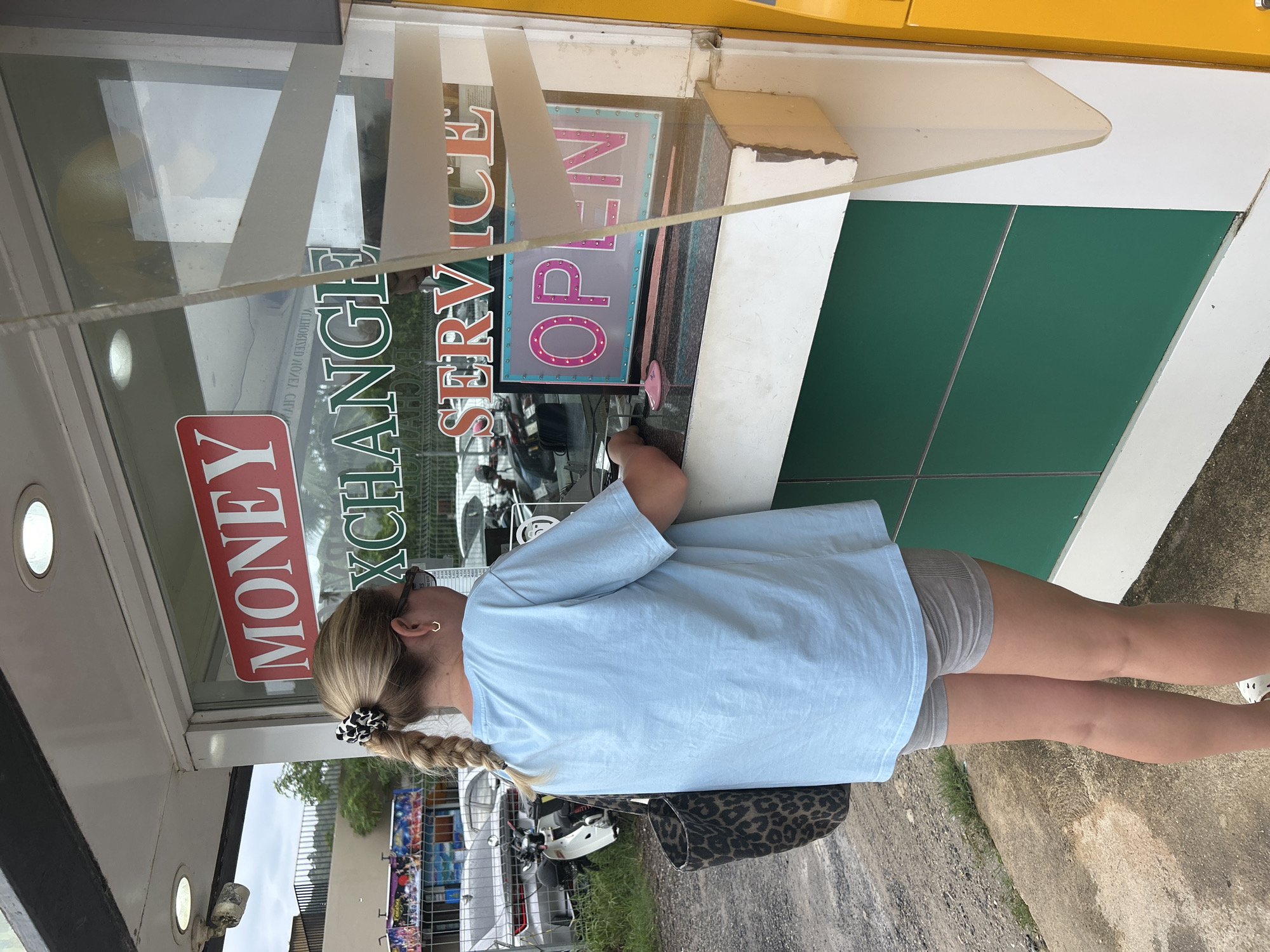 A woman with blonde hair styled in a braid, secured with a patterned scrunchie, is interacting with a Money Exchange service kiosk in Rawai, Thailand. She is wearing a light blue t-shirt and grey shorts and has a leopard print bag hanging from her shoulder. The kiosk features signage with the words "MONEY EXCHANGE SERVICE" and "OPEN". Outside the kiosk are parked vehicles and streetscape details.