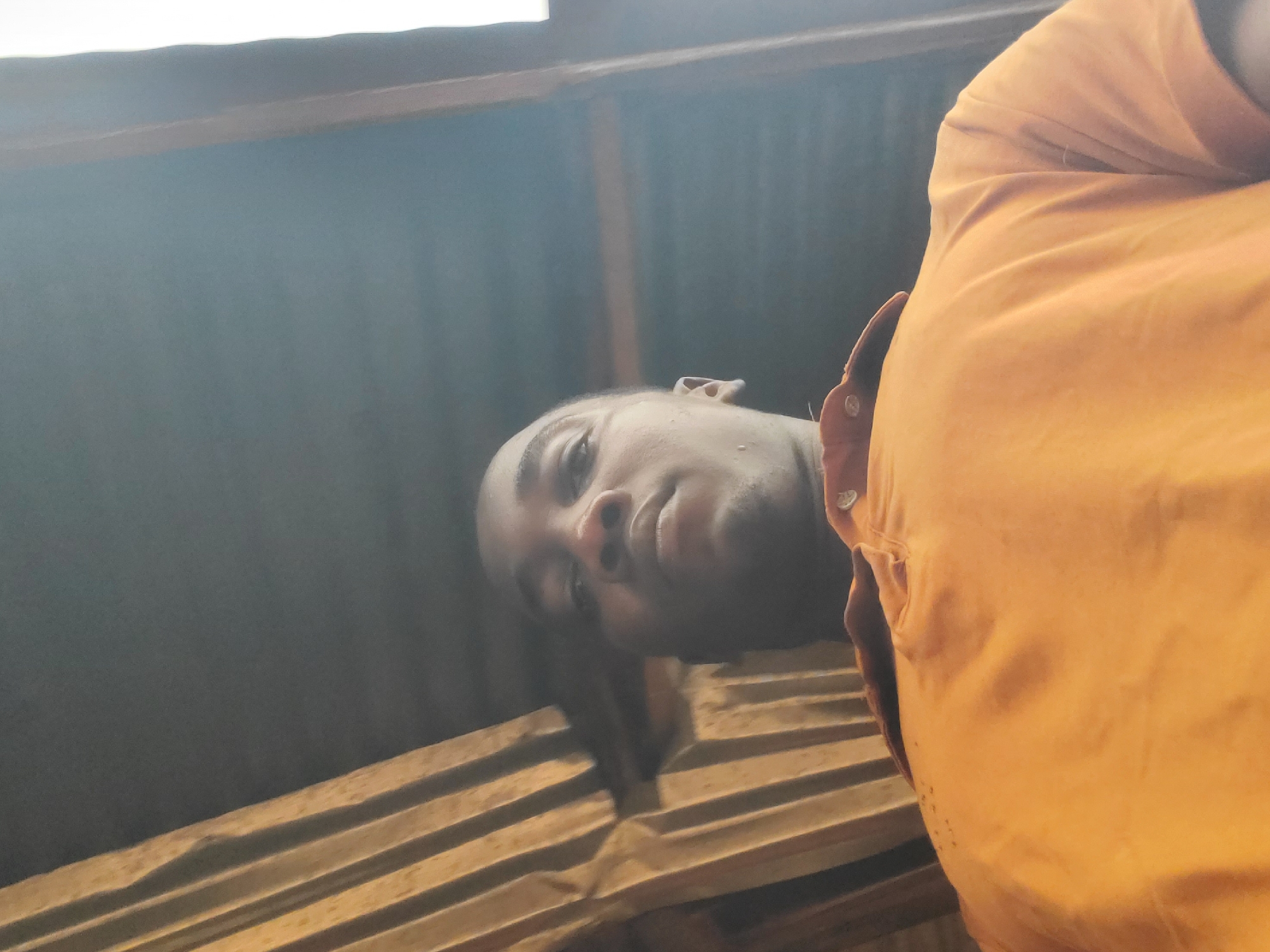 The image is a low-angle, selfie-style shot of a dark-skinned person wearing an orange shirt. The person's face is visible, and they are looking down at the camera. Behind the person, there is a gray corrugated metal structure, likely a roof or wall. Beneath the roof is wooden slats. The location is Ukwu-Abwa, Nigeria.