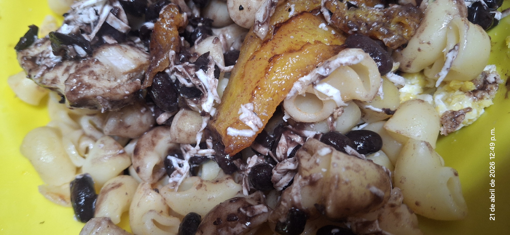 The image shows a close-up of a dish of food. The main ingredients appear to be elbow macaroni pasta, black beans, shredded cheese, slices of plantain and what may be diced chicken. Some of the pasta and chicken appear to be drizzled with a brown sauce. The food is presented in a yellow bowl. The image was taken on "21 de abril de 2026 12:49 p.m." according to the timestamp on the lower right. There is no indication of the location.