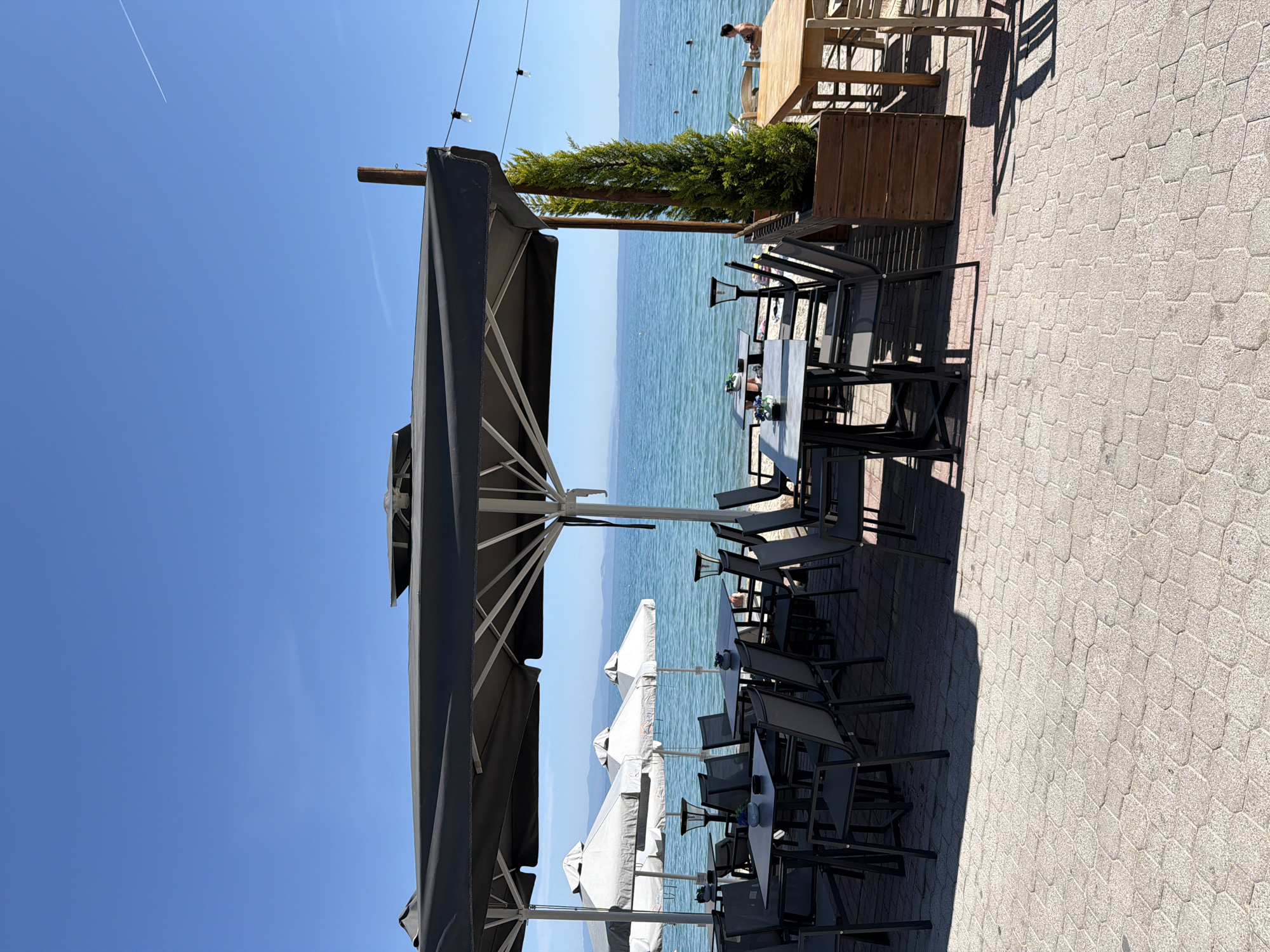 The image shows a restaurant patio in Pallini Municipal Unit, Greece. There are several tables and chairs set up, covered by large gray and white umbrellas. In the background is a body of water with a person standing on a dock or platform. There are trees in planters, and the ground is paved with gray patterned stones. The sky is blue with a contrail.