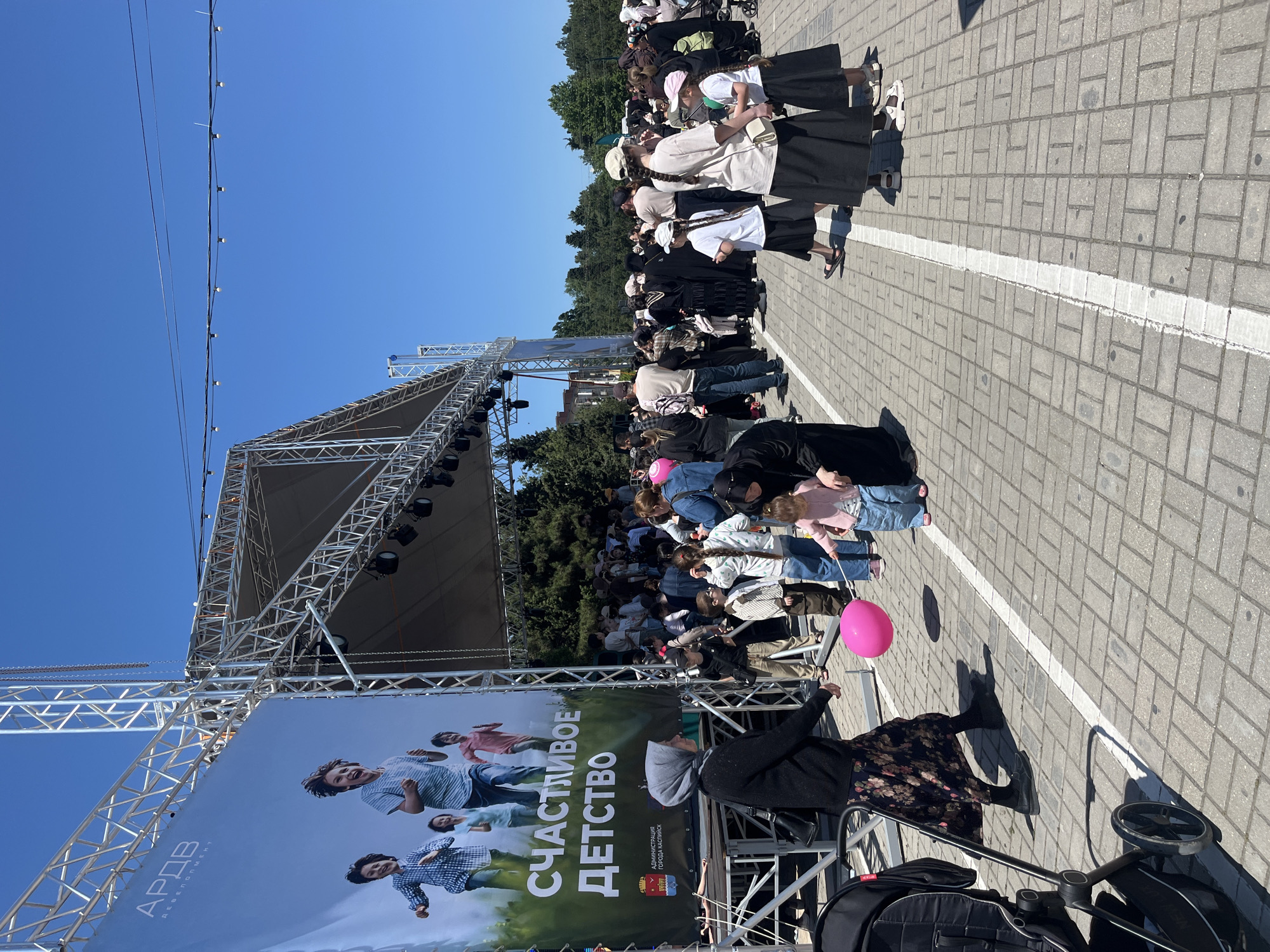 The image captures an outdoor event in Kaspiysk, Russia. A large stage dominates the scene, adorned with a banner that reads "СЧАСТЛИВОЕ ДЕТСТВО" (Happy Childhood) and includes the city's coat of arms. In front of the stage, a crowd of people, including women with covered hair and long skirts, are gathered, possibly anticipating or watching a performance. Many are families with young children. Some people are wearing jeans and casual shirts. A pink balloon floats near a woman standing with a stroller. The ground is paved with interlocking concrete blocks marked by a white line. Overhead, power lines stretch into the bright blue sky.