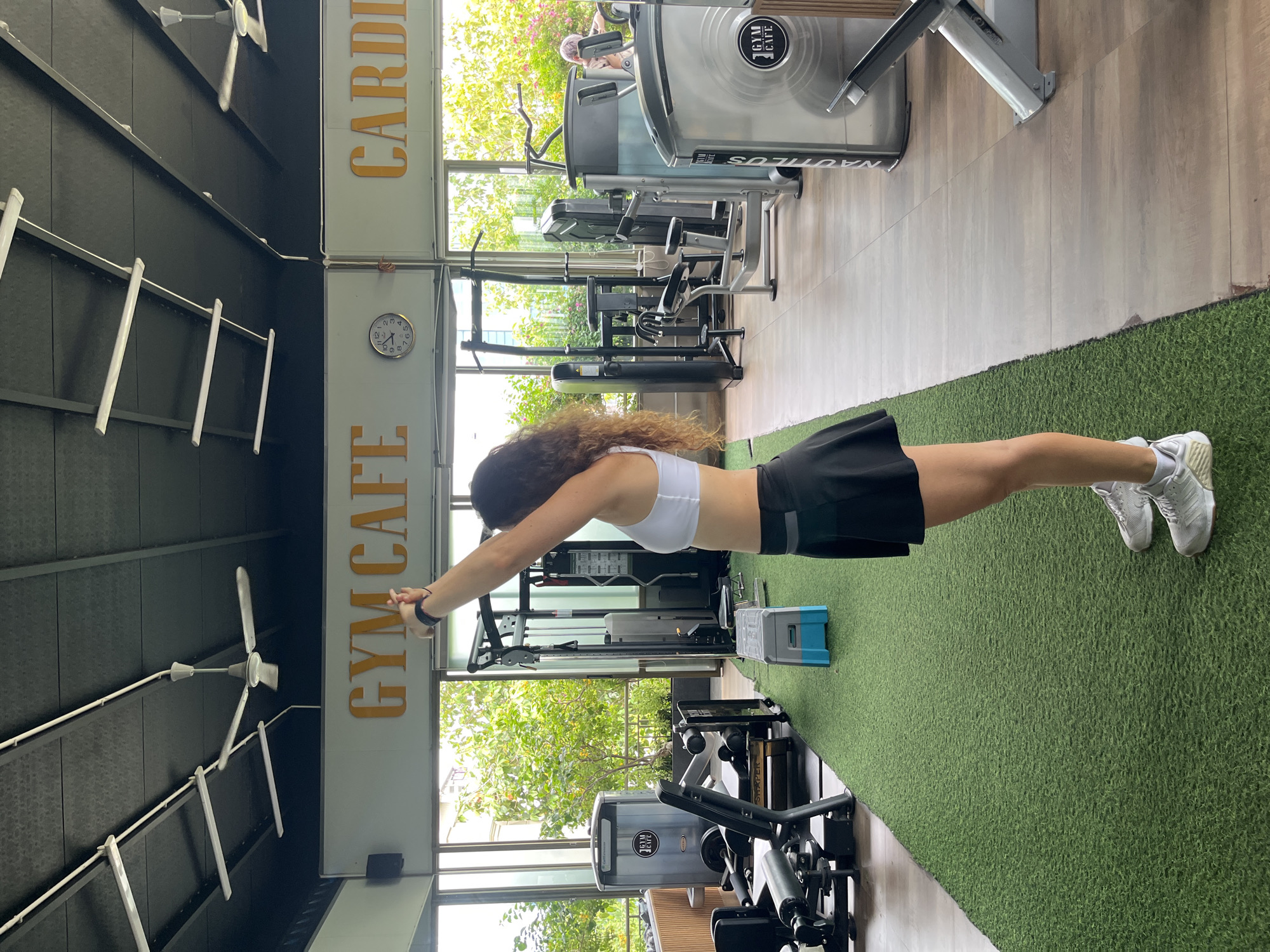 A woman with curly brown hair stands in a gym, facing left with her arms raised overhead. She is wearing a white sports bra, a black skirt, and white athletic shoes. In the background, gym equipment, including weight machines and cardio machines, is visible. The gym appears to be in Phan Thiet, Vietnam, indicated by the "GYM CAFE" signage and the tropical foliage visible through large windows. The floor is covered with artificial turf in the foreground and wood flooring in the background. There are ceiling fans and fluorescent lights on the ceiling.