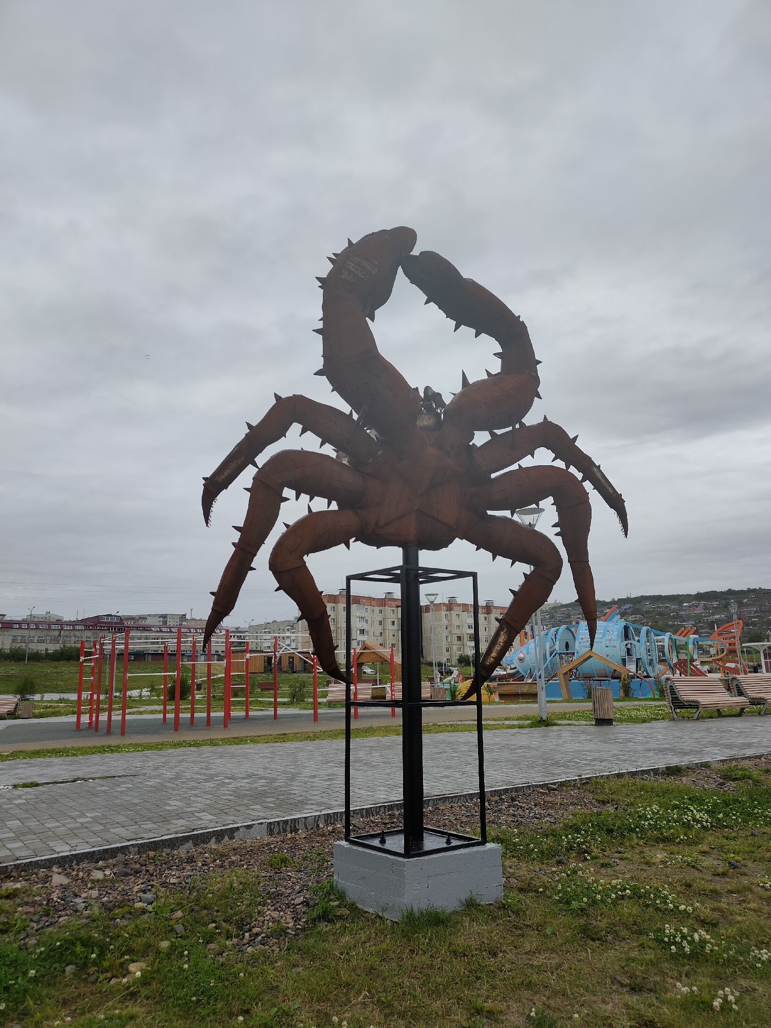 An outdoor sculpture of a large crab, made of rusted metal with sharp spikes protruding from its legs and claws, stands prominently in the foreground. The crab is mounted on a black metal pole supported by a grey concrete base.
Behind the sculpture, a paved walkway with grey cobblestones stretches across the scene, bordered by gravel and grass. In the mid-ground, a children's playground is visible with red horizontal bars for gymnastics, a large blue fish-shaped play structure, and wooden benches. Residential buildings, some with visible windows and balconies, are situated in the background, nestled against a hilly landscape under an overcast, cloudy sky. The overall environment appears to be a public outdoor space in the city of Magadan, Russia, likely a park or recreational area. No people are present in the image.