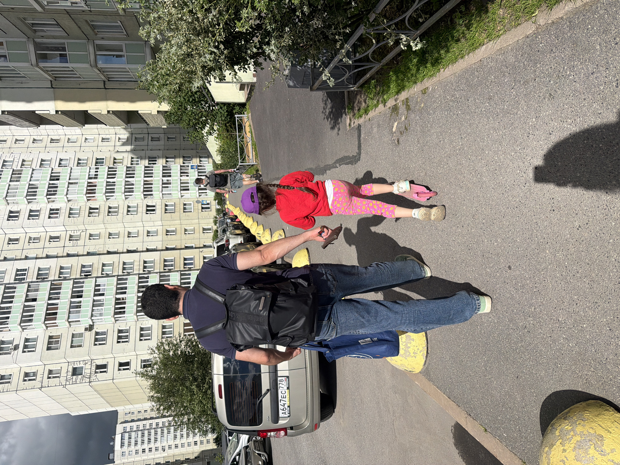 This is a slightly tilted, eye-level, medium shot of a young girl on a scooter and a man standing near her on a paved walkway. The girl is wearing a red long-sleeved shirt, pink leggings with a yellow pattern, and a purple baseball cap. She is facing away from the camera, with her hair in a braid, and is in motion on her pink scooter. The man is wearing a dark blue t-shirt, blue jeans, and a backpack. He is holding a dark object, possibly a phone, in his right hand.
The setting appears to be a residential area in Saint Petersburg, Russia, indicated by the background. Tall, multi-story apartment buildings with many windows dominate the left side of the frame. Trees and some greenery are visible along the walkway and in front of the buildings. A metal fence runs along the edge of the walkway. The ground is asphalt, with several large, yellow, bollard-like objects placed strategically. The lighting suggests it is daytime, likely sunny given the shadows. The overall mood seems casual and relaxed, with the girl engaged in outdoor play. A silver van with a Russian license plate (A647EC 778) is parked nearby. The text "CALVIN KLEIN JEANS" is visible on the man's backpack.