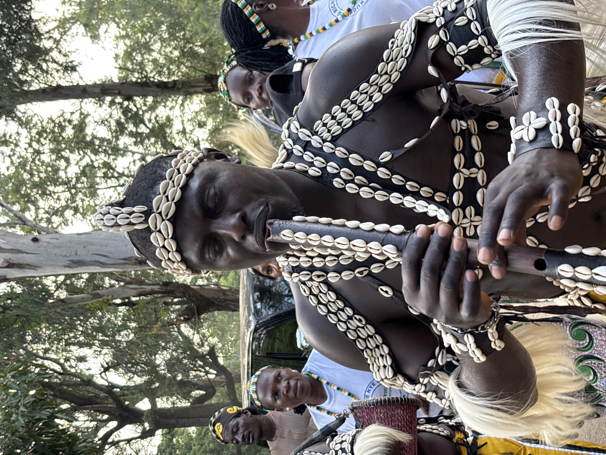 The image shows a group of people in traditional attire, likely participating in a cultural event or performance in Jos, Nigeria. The main subject is a man playing a wooden flute, adorned with necklaces and bracelets made of seashells. He is wearing a black vest decorated with multiple strands of seashells. His skin is dark, and he has short dark hair. His eyes are closed as he plays the instrument, suggesting deep concentration or immersion in the music.
Other individuals are visible in the background, also wearing traditional headbands with colorful beads and some form of seashell adornment. One person in the middle ground is wearing a white t-shirt with some text on it. To the left, another man is partially visible, also wearing a headband. A dark-colored vehicle is parked behind the central figure. The setting appears to be outdoors, with trees and foliage visible, indicating a natural or semi-natural environment, possibly a park or a rural area. The lighting suggests it might be daytime, perhaps late afternoon, given the soft light.
The overall mood seems to be one of cultural celebration and artistic expression. The intricate seashell decorations point to a rich cultural heritage, possibly related to traditional music or rituals. The people's attire and the presence of the flute are strong indicators of a cultural performance or ceremony. The image captures a moment of musical performance within a communal gathering.