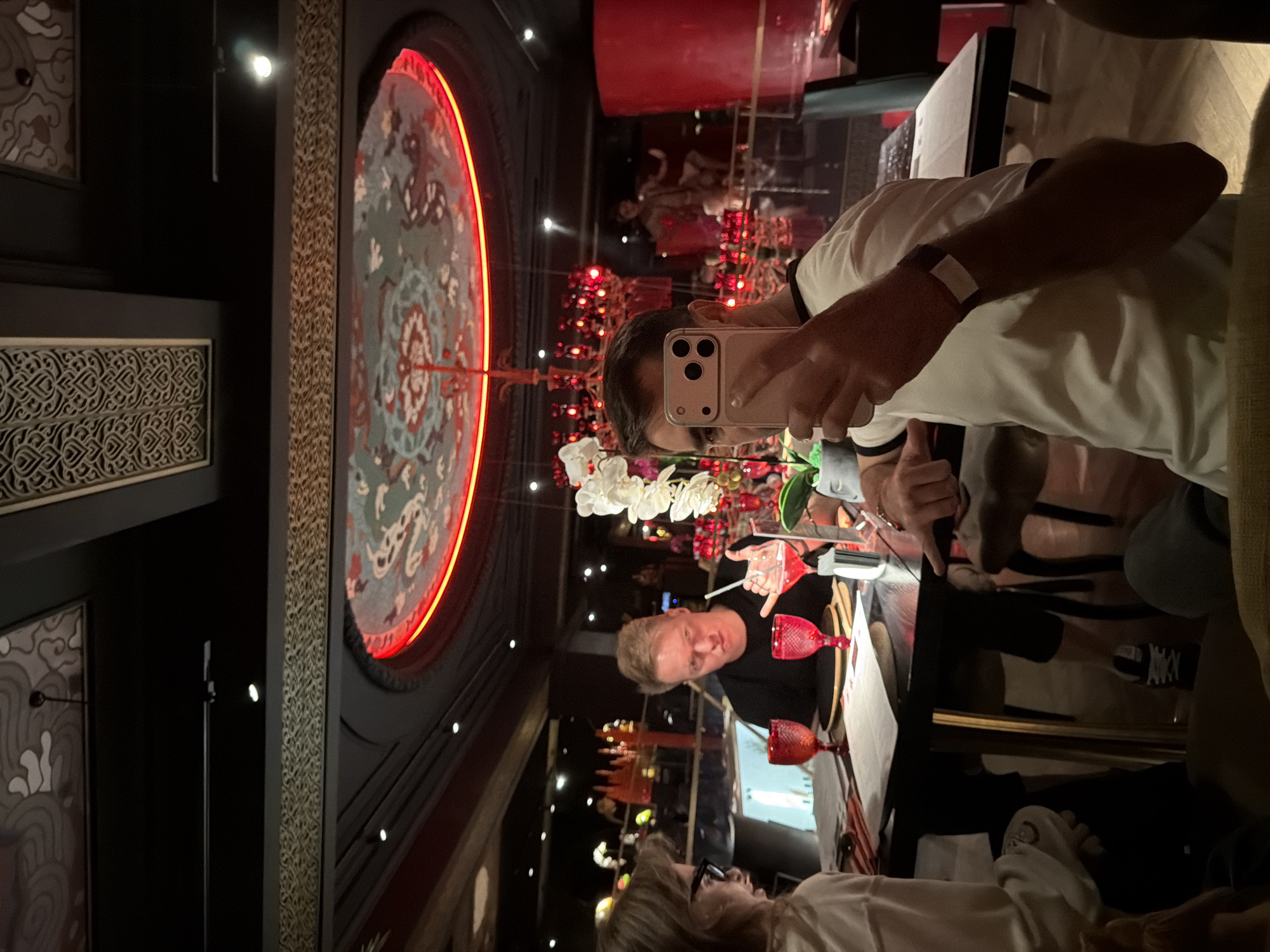 The image captures an interior scene in what appears to be an upscale restaurant or lounge, possibly located in Moscow, Russia, given the ornate decor. A large, circular, decorative panel with an intricate design and illuminated with red neon lighting dominates the left side of the frame.
Several individuals are present. In the foreground, a person is taking a selfie with a smartphone, their face partially obscured by the device. Another person with blond hair is seated at the table, facing the camera. Further down the table, another individual is visible, wearing glasses.
The scene is set at a dining table with several decorative red glasses. Floral arrangements add to the ambiance. The background suggests a bustling restaurant environment with other patrons and decorative elements. The image is a snapshot of an evening social gathering within a luxurious setting.