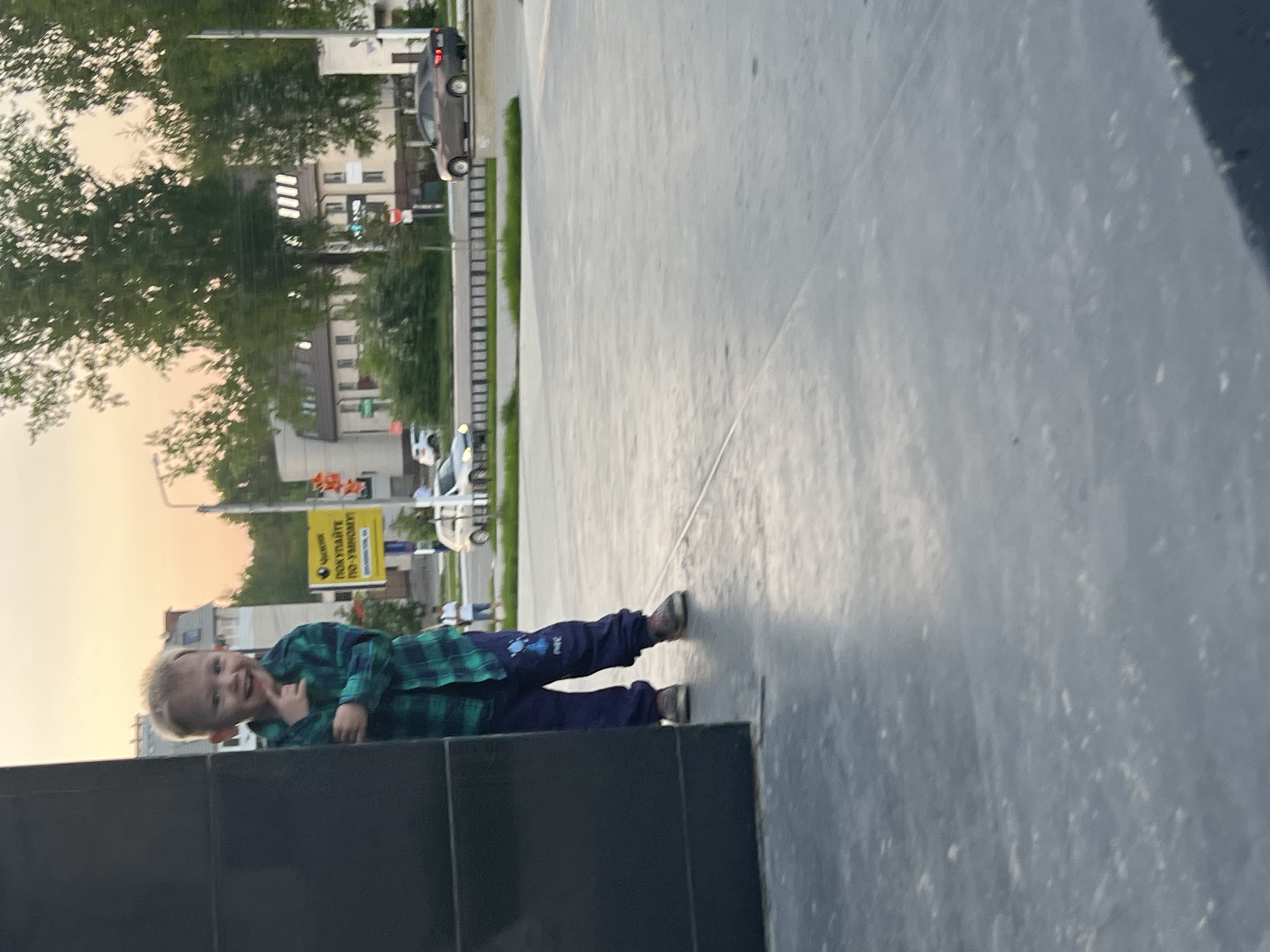 A young child is lying on a gray concrete surface near a low, black wall. The child wears a green plaid shirt and dark pants. A road with buildings and trees is visible in the background. A yellow sign with Cyrillic text is also visible. Based on the available information and visual cues, this image seems to be in the city of Chaikovsky, Russia.