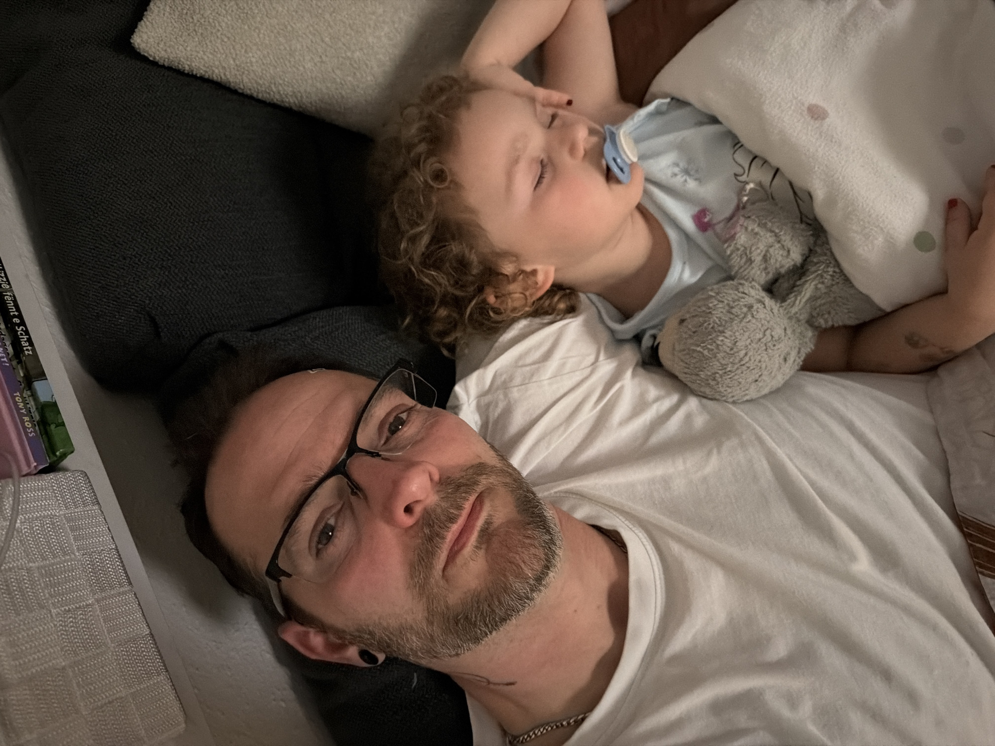 A close-up shot shows a man and a child lying down, presumably indoors. The man, wearing glasses and a white t-shirt, is positioned at the bottom of the frame. The child, sleeping with a pacifier in their mouth, lies above the man, partially covered by a white blanket and clutching a stuffed rabbit. Part of a shelf with books on it can be seen in the top left. The location is Trassem, Germany.