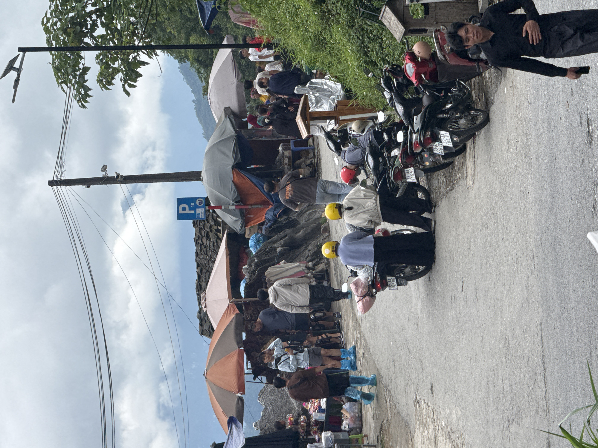 A ground-level, medium-shot image captures a street scene in Yên Minh Commune, Vietnam. The viewpoint is along a roadside, where people are engaged in street commerce under several umbrellas, and motorcycles are parked. The photograph is taken at an angle, with the foreground slightly higher than the background.
On the right, a man is lying on the ground beside the parked motorcycles, holding a cell phone in his right hand. Behind him are several motorcycles, their license plates partially visible. Several people are near the vehicles. The license plates have Vietnamese writing.
On the left side of the scene, under the shade of a large orange umbrella, people are observed shopping at a roadside market. Many of them are wearing blue plastic shoe covers, indicating potential muddy conditions. Further in the distance, more people are congregated around tables and vendors. An old parking sign, “P 18,” is attached to the side of a stone wall. Overhead, power lines stretch across the sky, running diagonally from the left side of the scene toward the background.
The environment appears to be a mountainous region, as evidenced by the rising terrain and lush greenery in the background. The sky is filled with gray clouds, suggesting overcast weather.