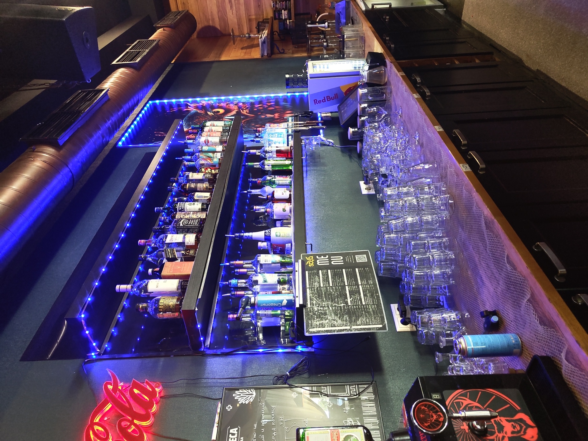This image shows a bar interior, likely located in Zhukovsky, Russia, given the text on the menu board. The bar is dimly lit, with prominent blue LED lighting illuminating shelves of liquor bottles and rows of glassware. A neon red Coca-Cola sign is visible on the left wall. The bar counter is covered with a white mesh-like material and features stacks of clean drinking glasses, a beverage dispenser labeled "Jägermeister", and a blue CO2 canister. Behind the counter, shelves are stocked with various alcoholic beverages, including bottles of whiskey, vodka, and liqueurs. A large menu board is displayed on the wall. The overall atmosphere suggests a modern and well-equipped bar or lounge.