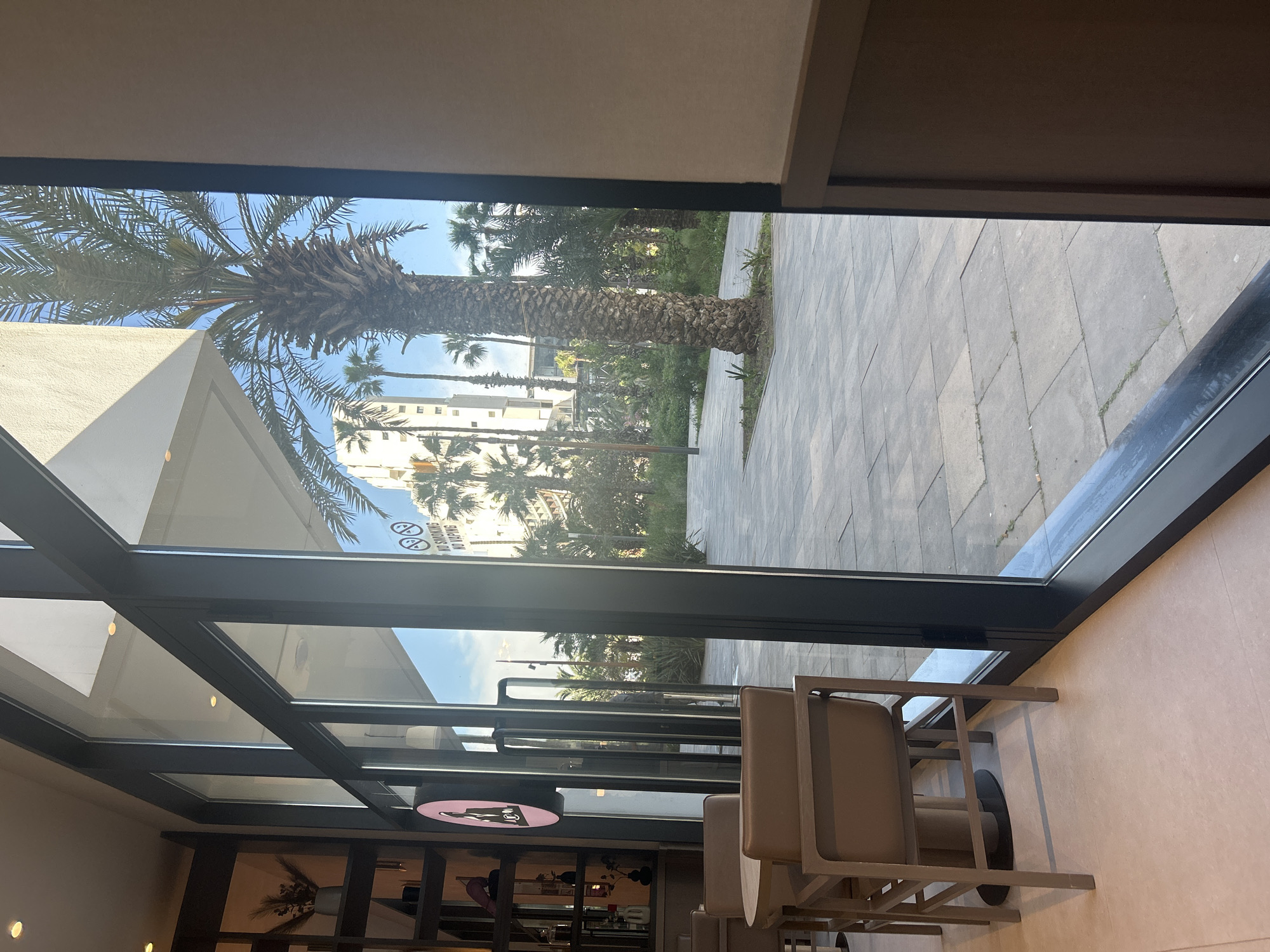 The photo depicts a view from inside a building, likely a cafe or restaurant, looking out onto a cityscape in Casablanca, Morocco. A large palm tree is visible outside the window, along with other vegetation and buildings. The foreground shows indoor furniture, including chairs and tables. There is a glass window with the reflection of outside scenery. The overall environment suggests an upscale establishment in a city setting. The photo is taken at an angle, which makes the vertical scene seem sideways.