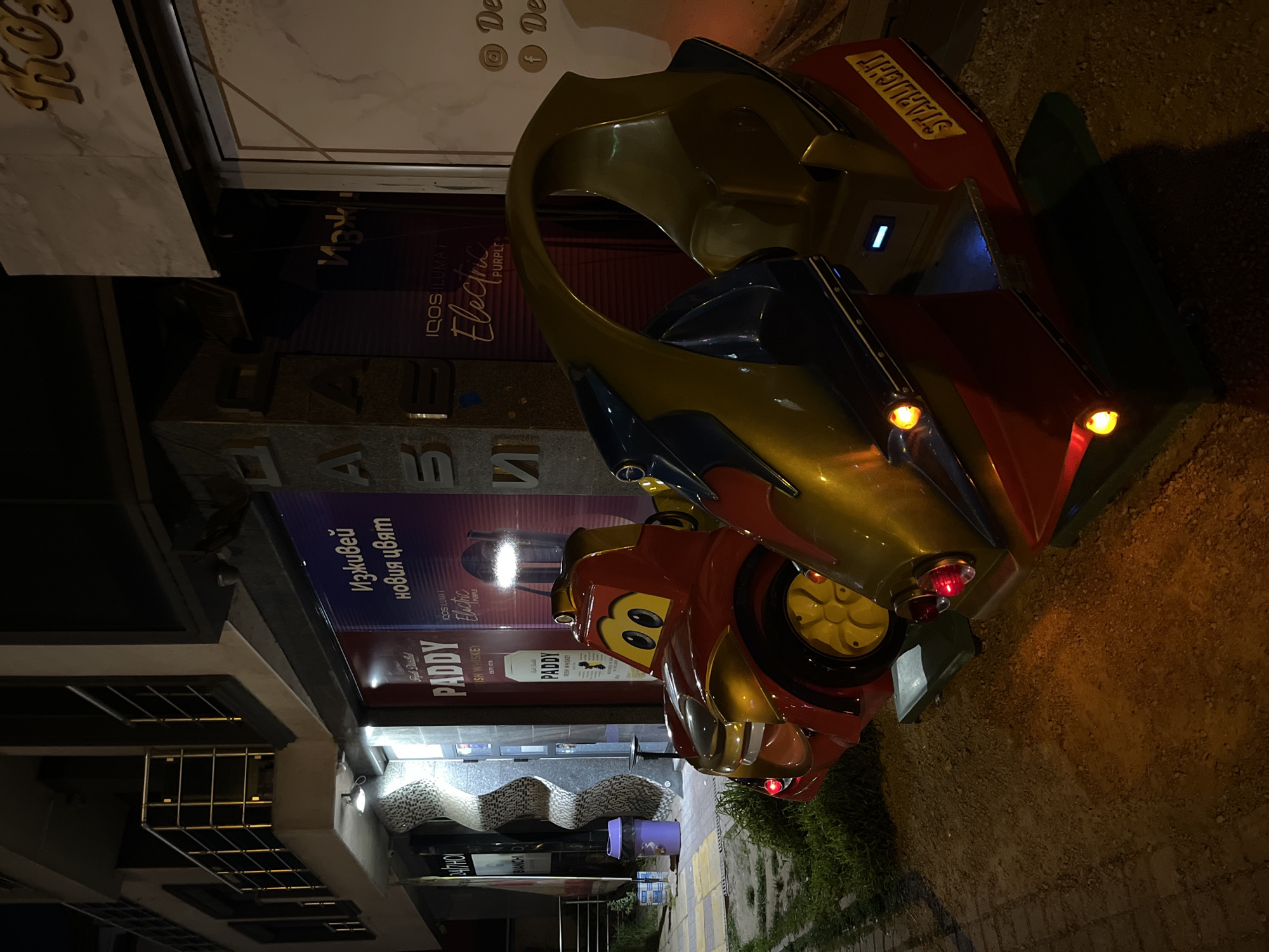 The image captures a nighttime street scene in what appears to be an urban commercial area, likely Sofia, Bulgaria, given the Cyrillic text on various signs. The central focus is a vibrant, coin-operated children's ride, resembling a futuristic car or space vehicle, predominantly red and gold with yellow wheels and details. It features a cartoonish face on the front and illuminated headlights and taillights. The word "STARLIGHT" is visible in yellow lettering on its side.
The ride is situated on a paved surface next to a small patch of grass, in front of several commercial establishments. The buildings are dark but illuminated by various signs and internal lights. Prominent advertisements include "IQOS ILUMAT" and "Electric PURPLE," alongside the Cyrillic phrase "Изживей новия цвят," which translates to "Experience the new color." Multiple banners for "PADDY IRISH WHISKEY" are also visible. Other partial texts include "24/7HO," "BIANCHI," and what appears to be "Grypal Dinilld" or a similar Cyrillic phrase. Golden stylized lettering, possibly "KHO," adorns a white facade.
No people are present, indicating a quiet late evening or nighttime setting. A purple trash can and a distinctive wavy-patterned bench or decorative wall element are also visible near the storefronts. The scene is static, lit by artificial light sources.