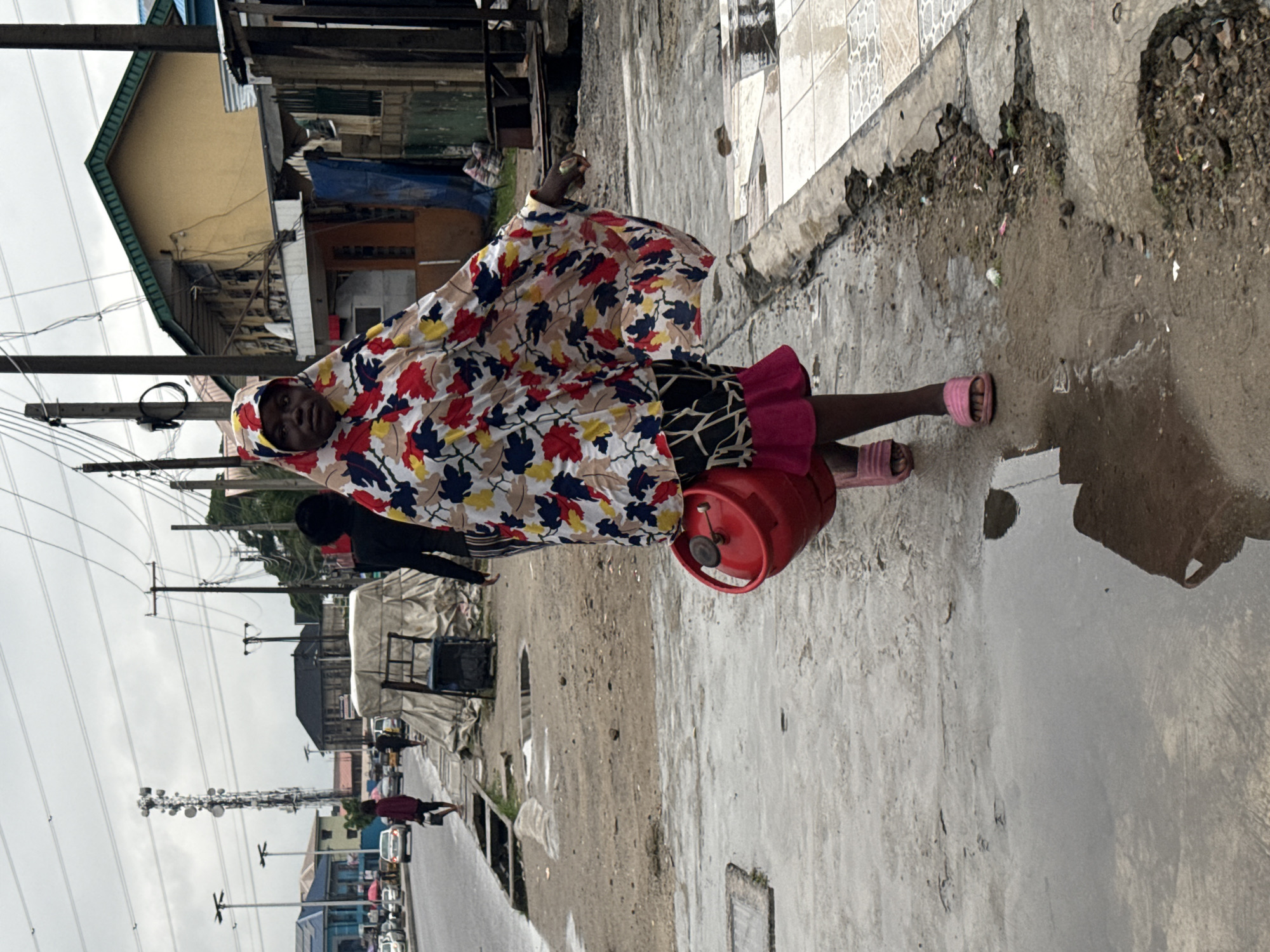 A woman walks along a wet street in Ado, Nigeria. She wears a patterned, hooded garment and pink sandals, carrying a red gas cylinder under her arm. Buildings and utility poles line the street. Cars are visible in the distance. The sky is overcast.